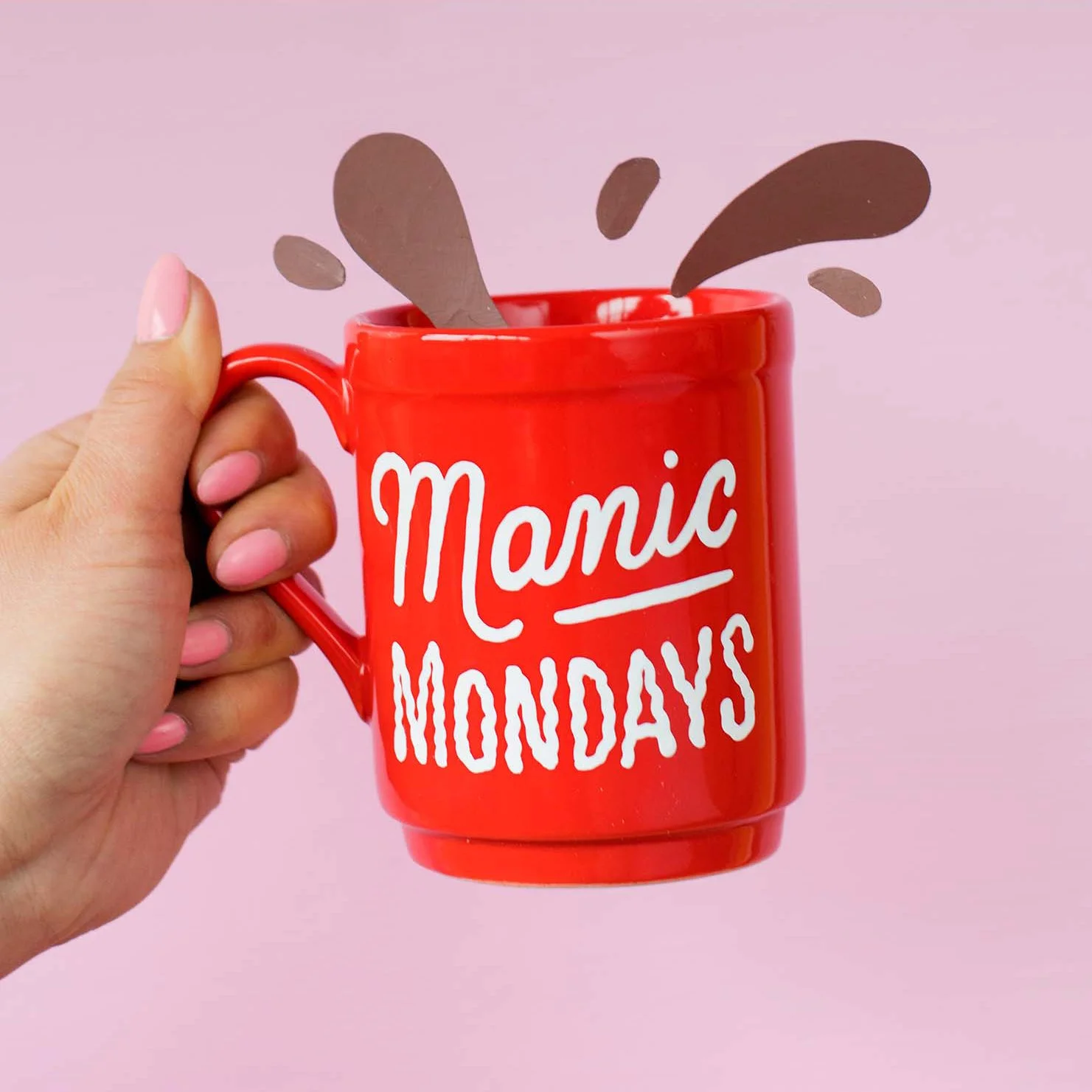 A Manicured hand holds a bright red coffee mug that says "Manic Mondays." Splashes made from paper emerge from the top of the mug.