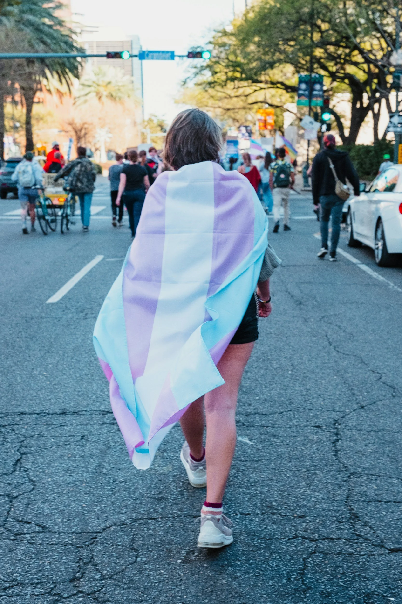Person walking across a city street with a LGBTQ+ pride flag draped over their shoulders. Other pedestrians are visible in the background, some with backpacks, a bicycle, and umbrellas. There are trees and traffic signals in the scene.