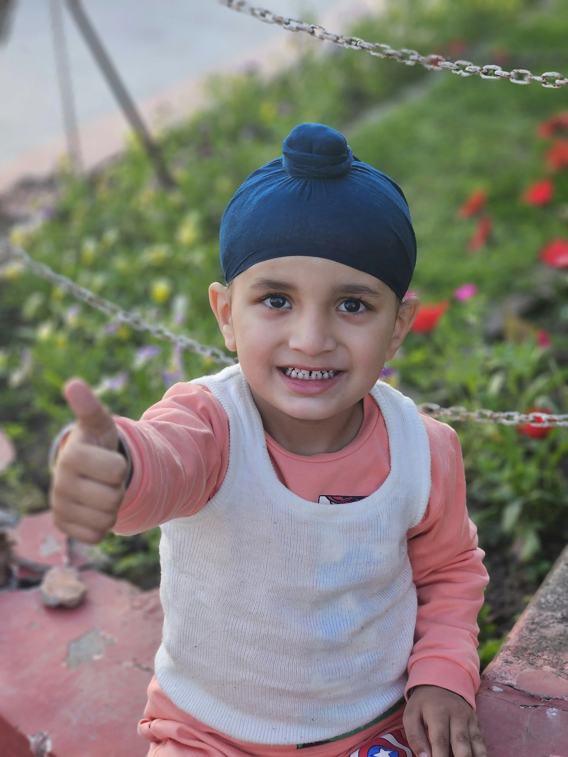 A young child wearing a blue turban and a pastel pink and white jacket is smiling at the camera, giving a thumbs-up, outdoors with blurred greenery in the background.