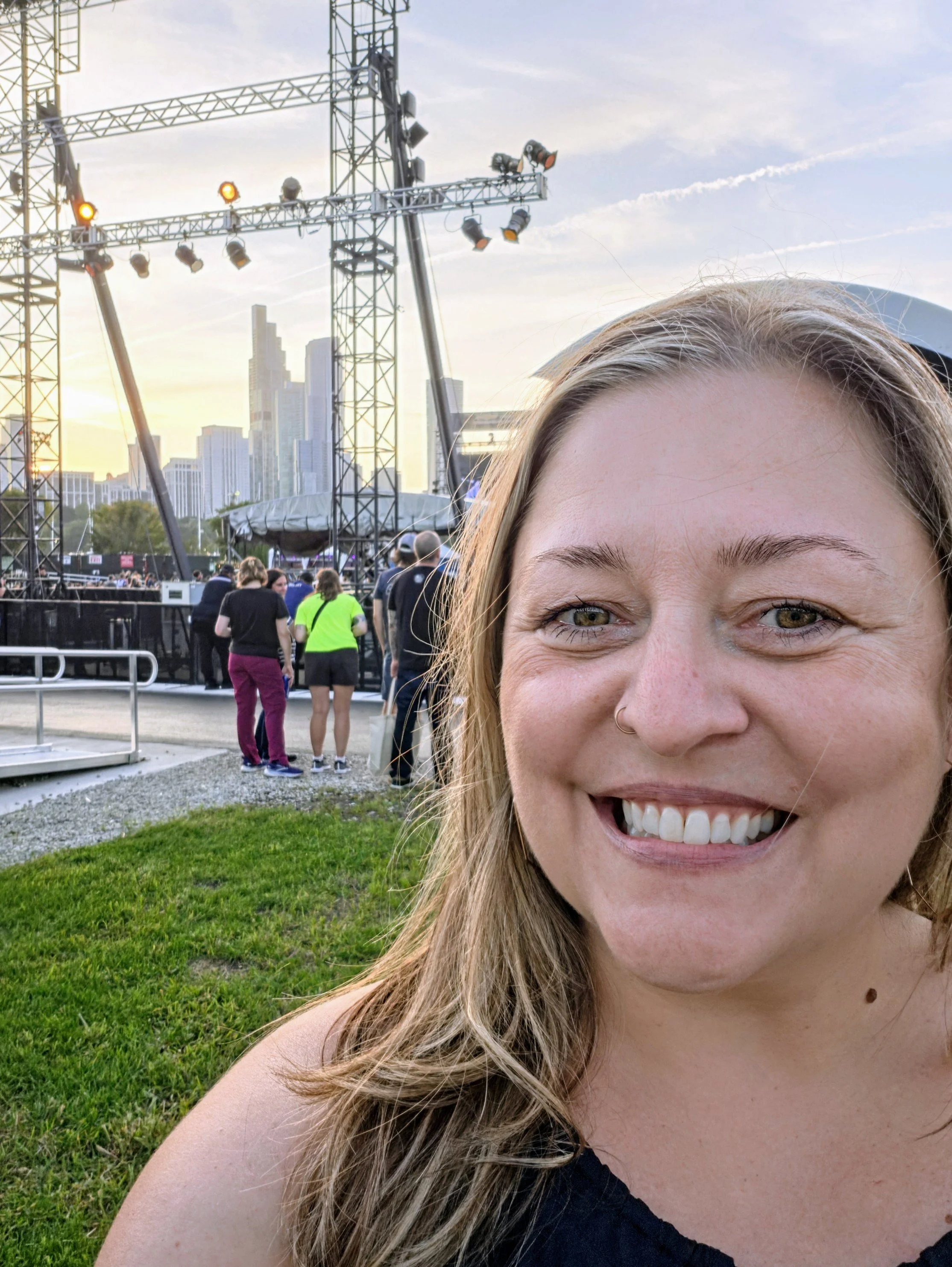 Close-up of a woman with blonde hair smiling, at an outdoor event with stage lights and a city skyline in the background.