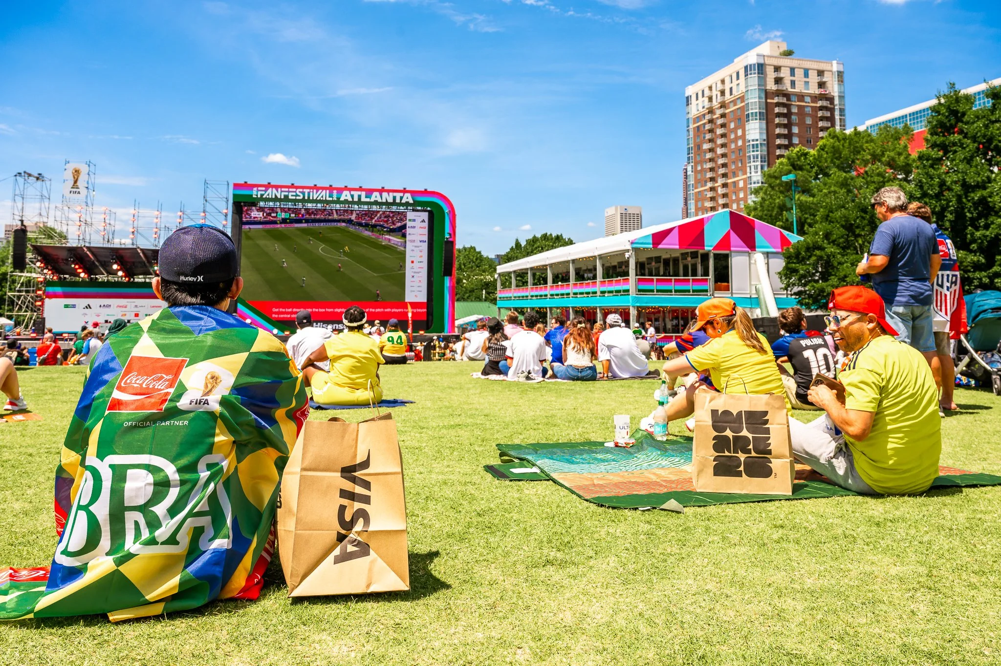 FIFA and Visa branded merchandise bags reading "We Are 26" and "Visa" rest together as fans watch the match with family and friends at FIFA Fan Fest Atlanta, highlighting the event's presenting sponsors.