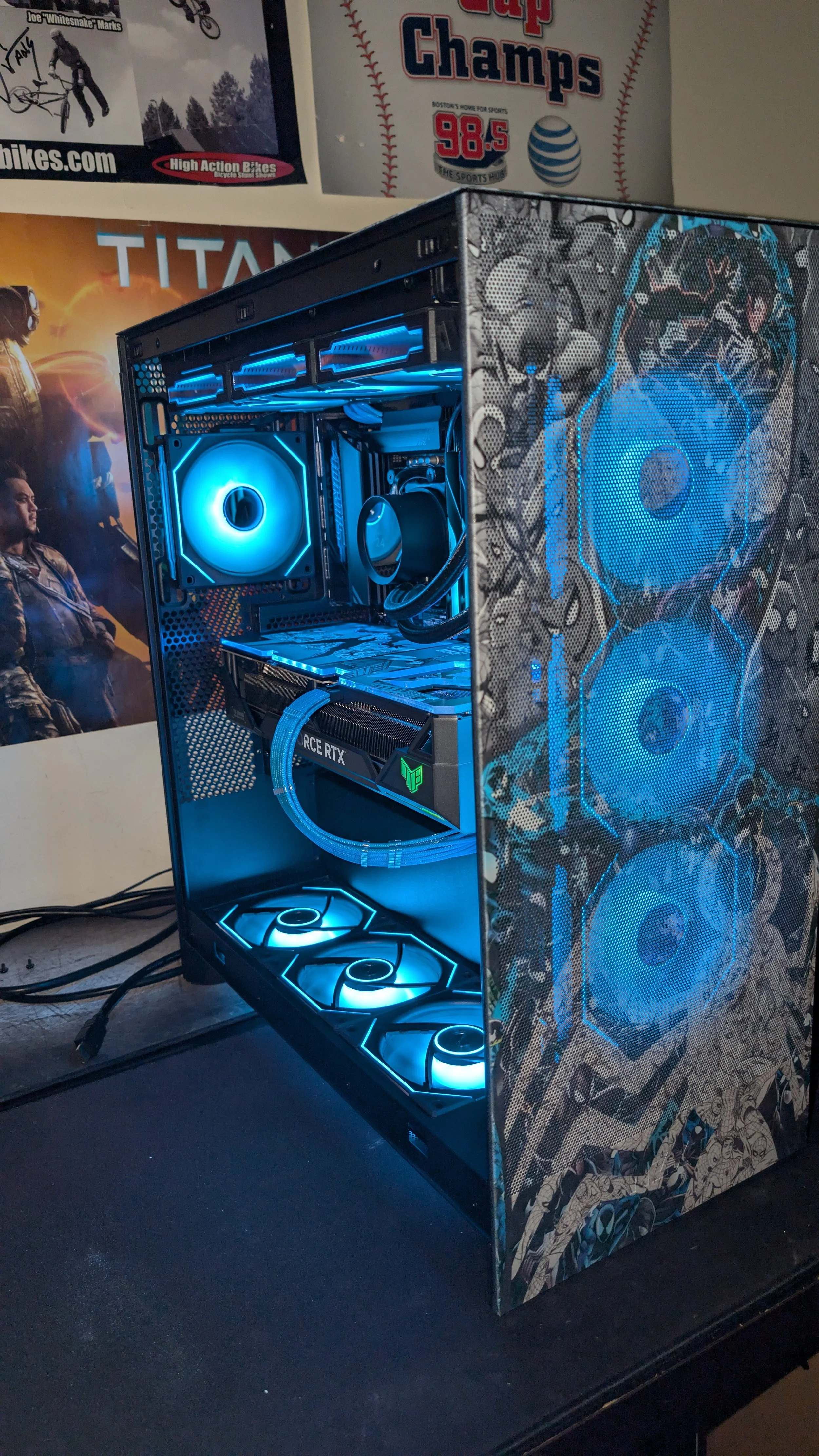 A custom-built gaming computer with illuminated blue LED fans and a water cooling system inside a decorative case on a desk, with posters and banners on the wall.