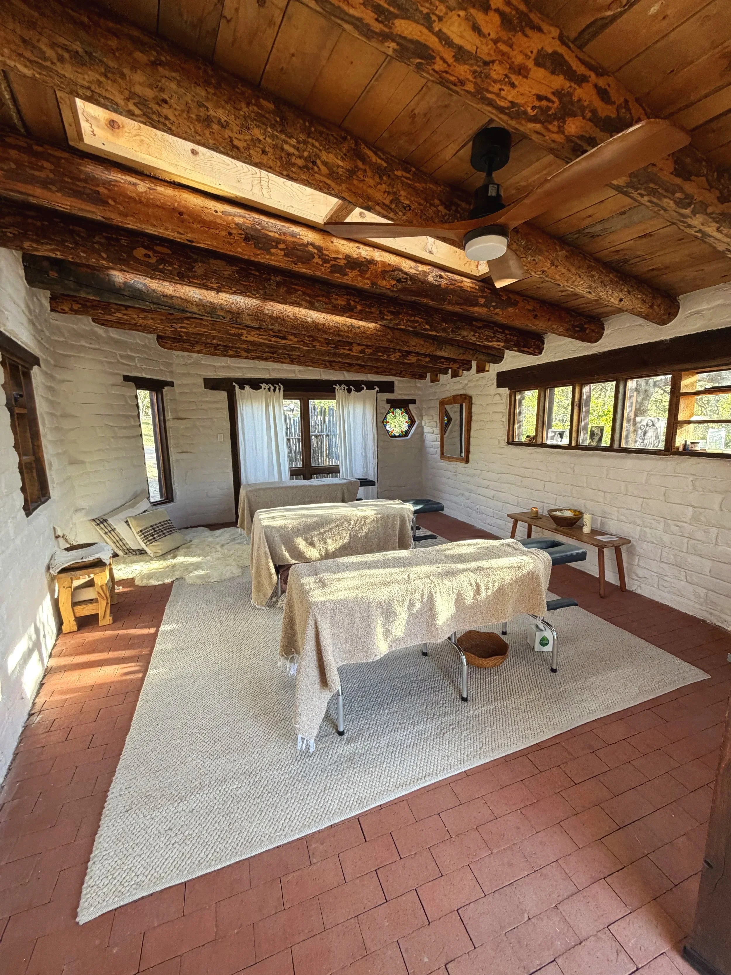 A cozy living room with white brick walls and wooden ceiling beams. There are large windows with white curtains, and several tables covered with beige cloths. A ceiling fan hangs from the wooden ceiling, and the floor is made of reddish bricks. Some framed artwork and photographs are visible on the walls.