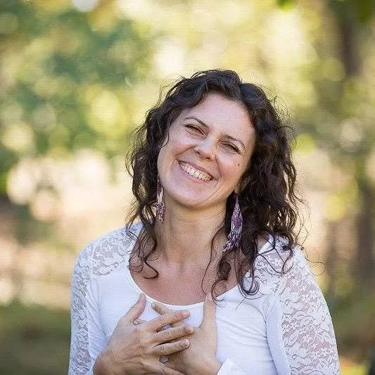 A woman with curly dark hair smiling outdoors, holding her hand to her chest, in a natural setting with trees and sunlight in the background.