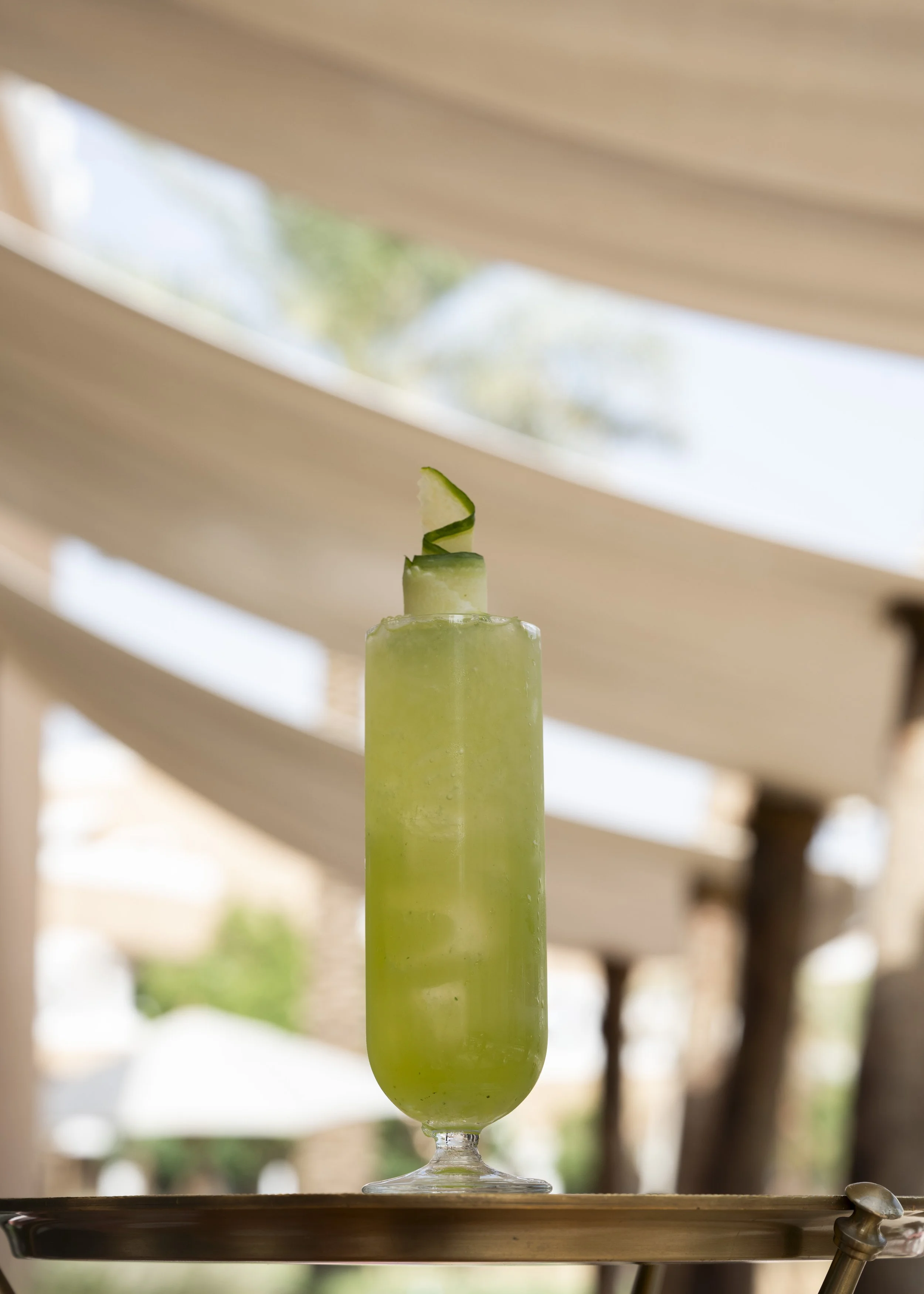 Green cucumber cocktail garnished with a cucumber peel curl in a tall glass on a tray, with blurred outdoor background.