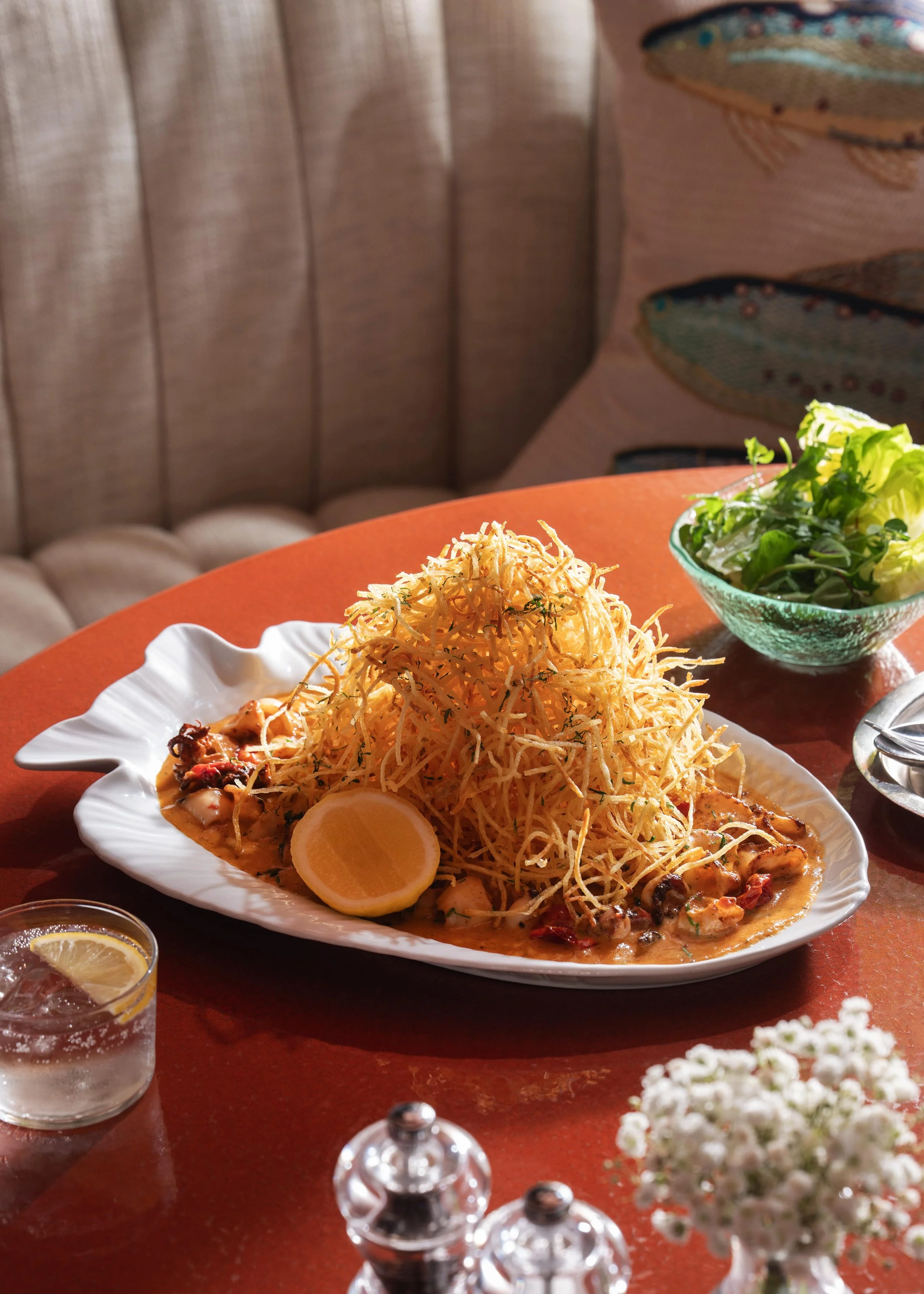 A seafood dish topped with crispy fried potato strings, served with a lemon wedge, along with a side salad in a glass bowl, on a red table.