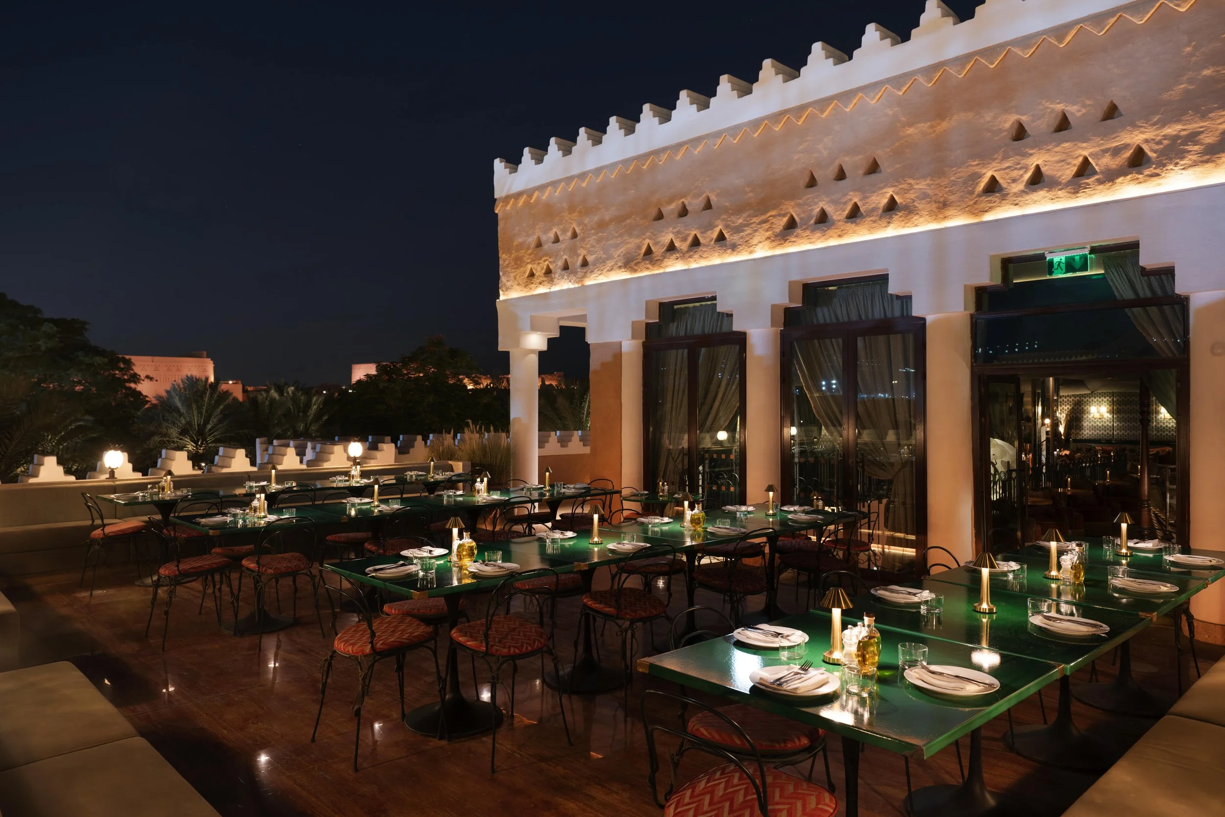 Outdoor rooftop restaurant at night with tables set for dining, large windows, decorative lighting, and a view of the dark sky.