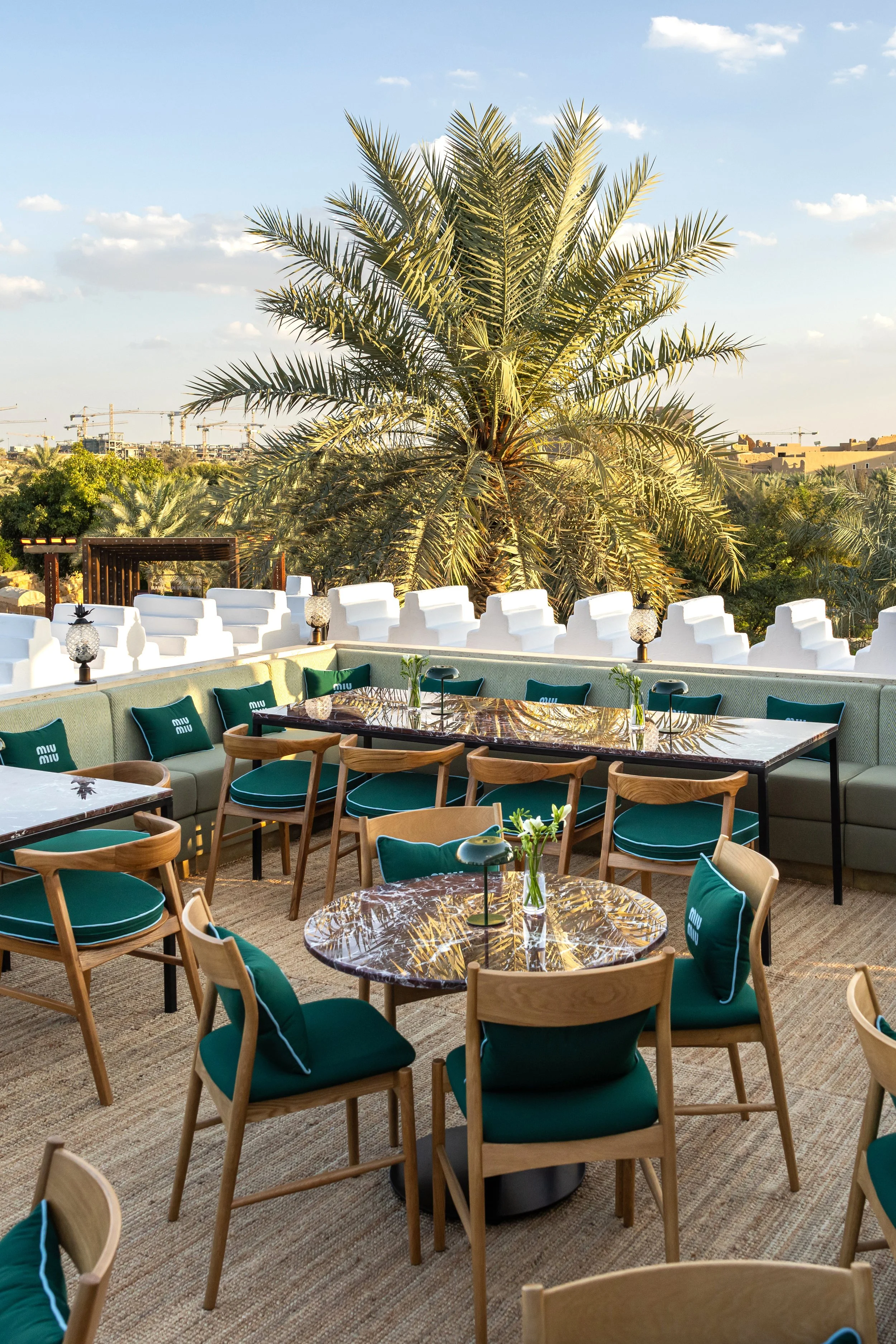Outdoor rooftop dining area with tables, chairs, green cushions, and large palm tree in the background under a clear sky.