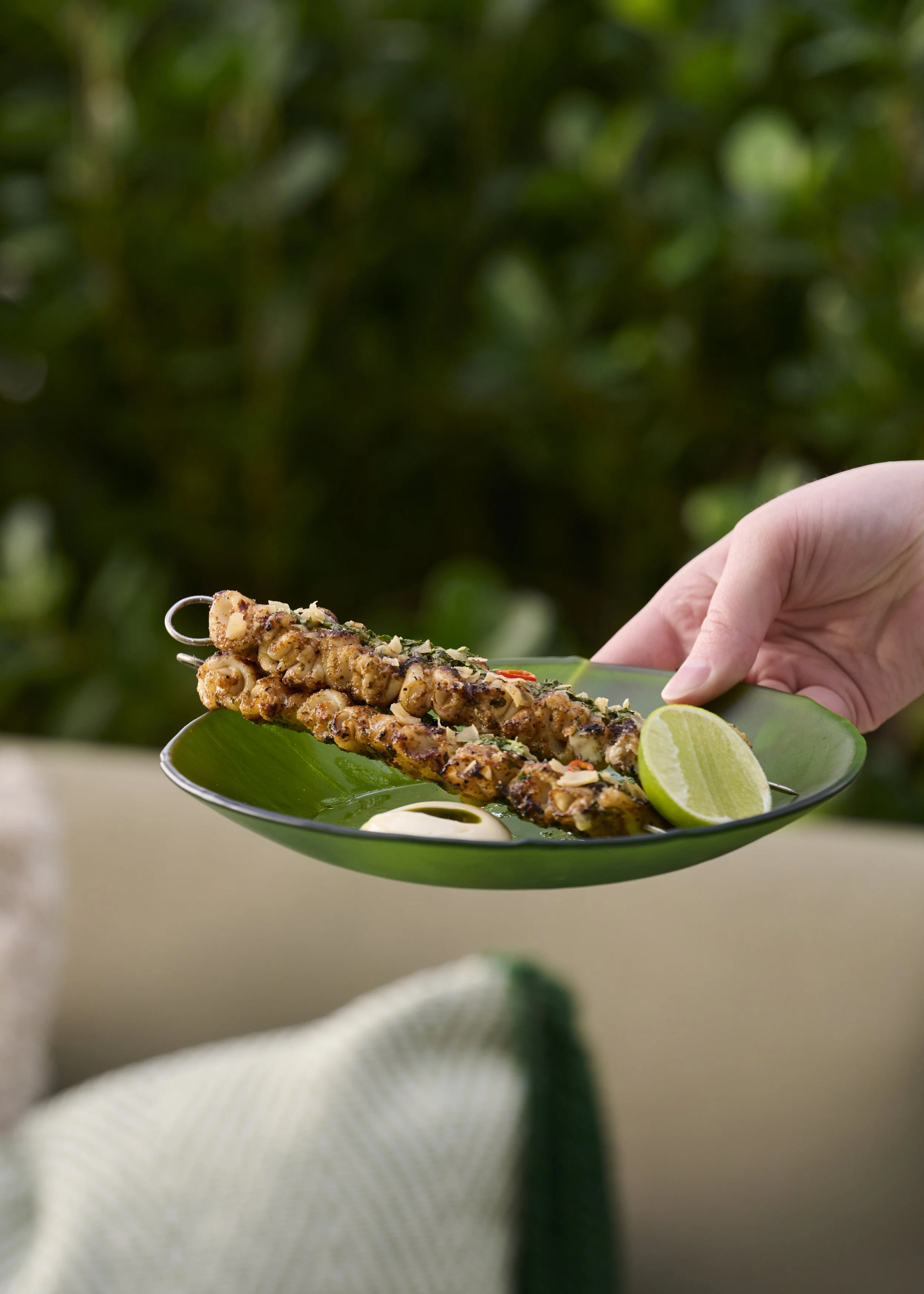 Grilled skewered seafood served on a green plate with lime and sauce, held by a hand with a blurred outdoor background.