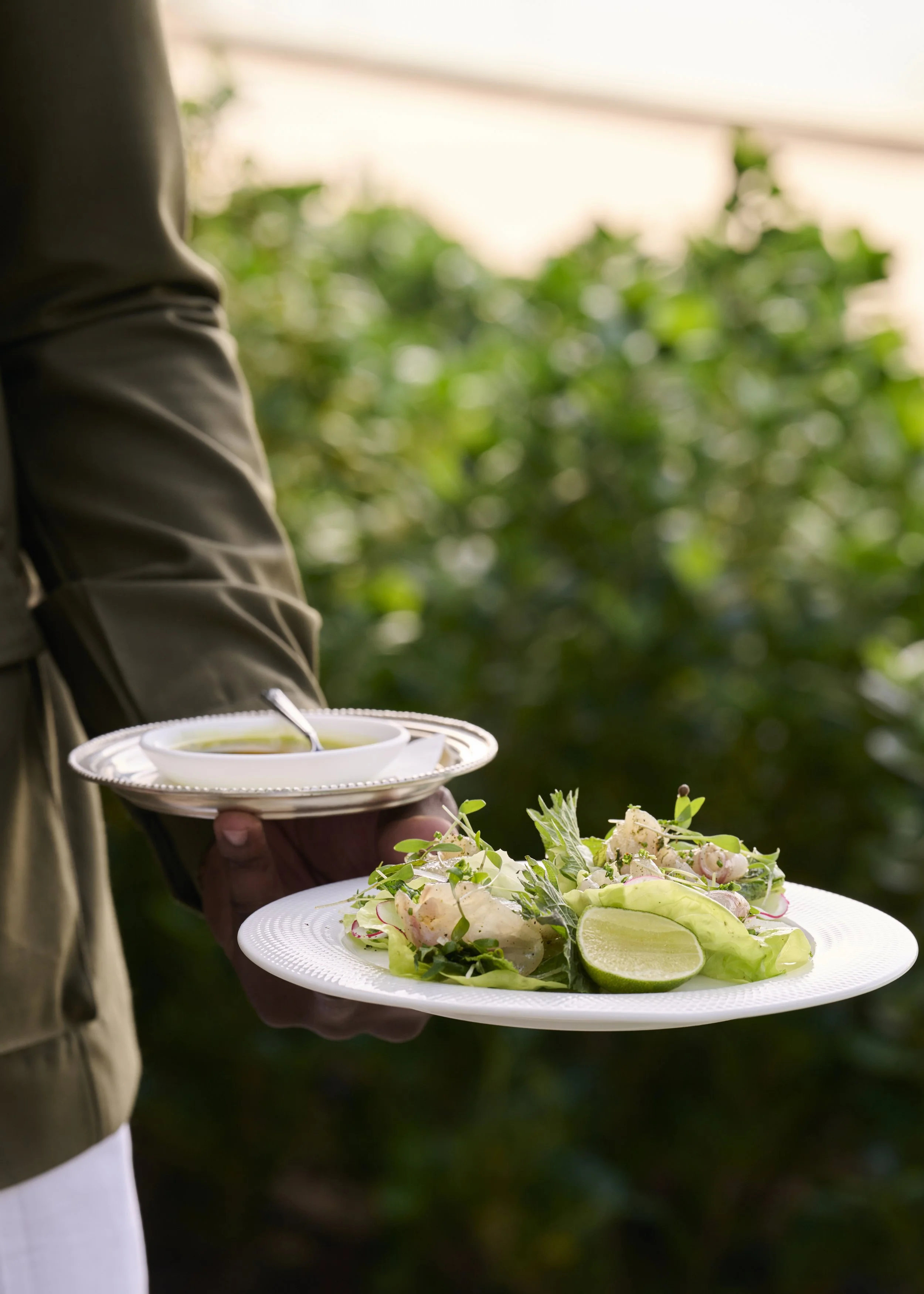 A person in a brown jacket holding a plate with a lime wedge, greens, and seafood salad, and a small bowl of dressing or sauce in an outdoor setting with greenery in the background.