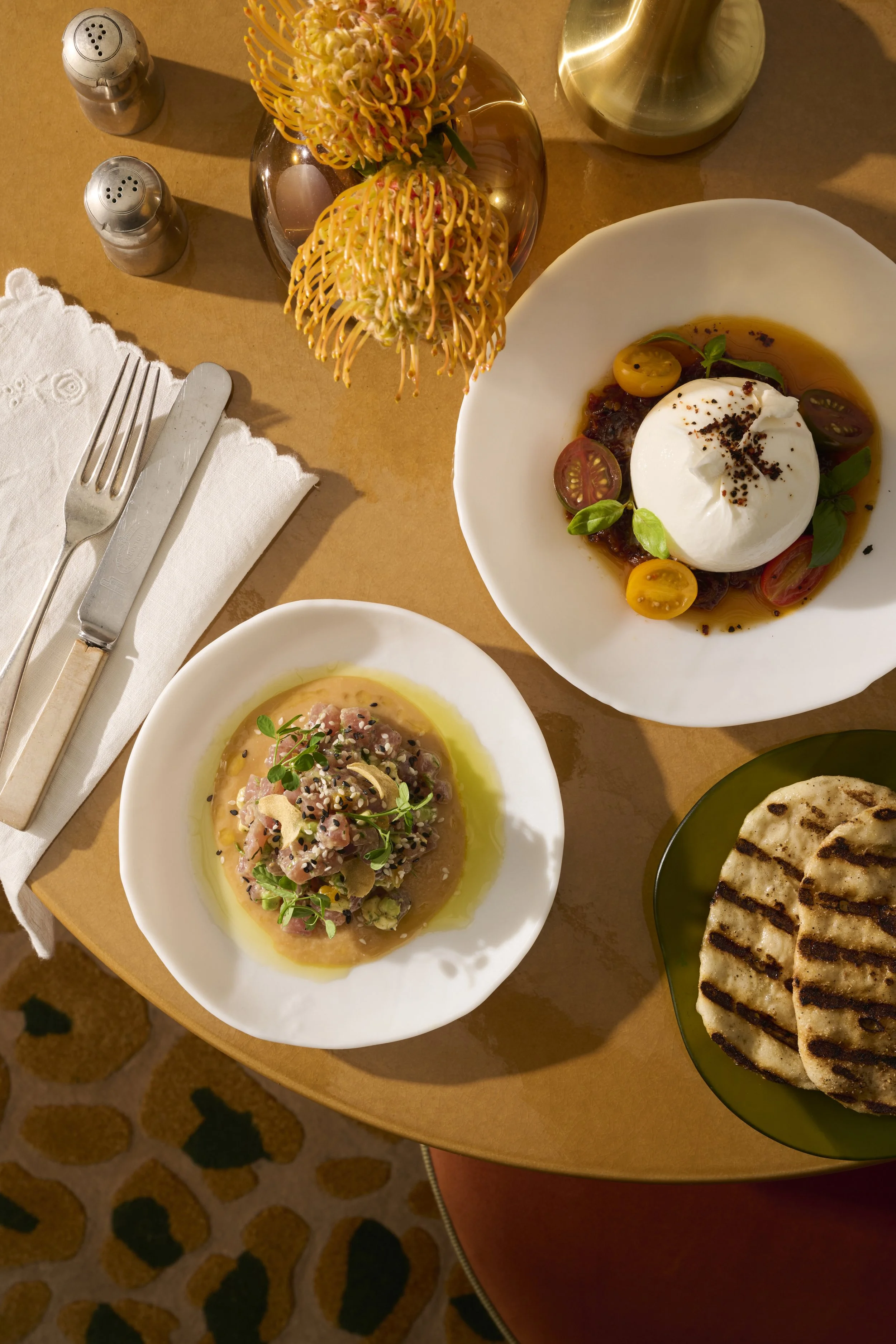 A table with two plates of food, a glass of water, salt and pepper shakers, a vase with flowers, and grilled flatbread on a small green plate. One dish is a burrata cheese with cherry tomatoes and basil, and the other is a beef tartare garnished with