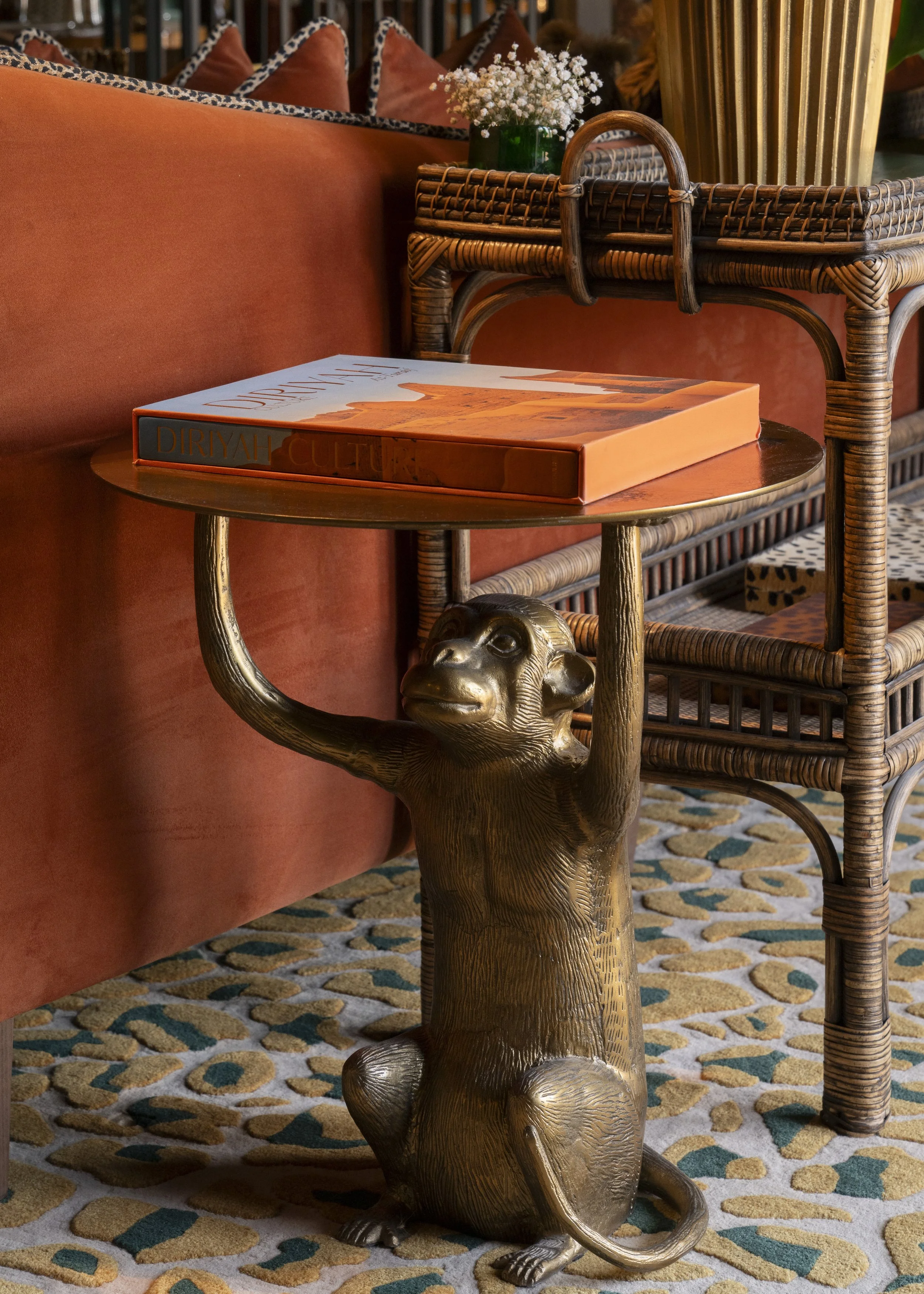 A decorative side table with a brass monkey sculpture as the base, holding a round wooden top. The table is next to a coral-colored sofa with patterned pillows and a wicker chair. A green vase with white flowers is on a surface behind the table.