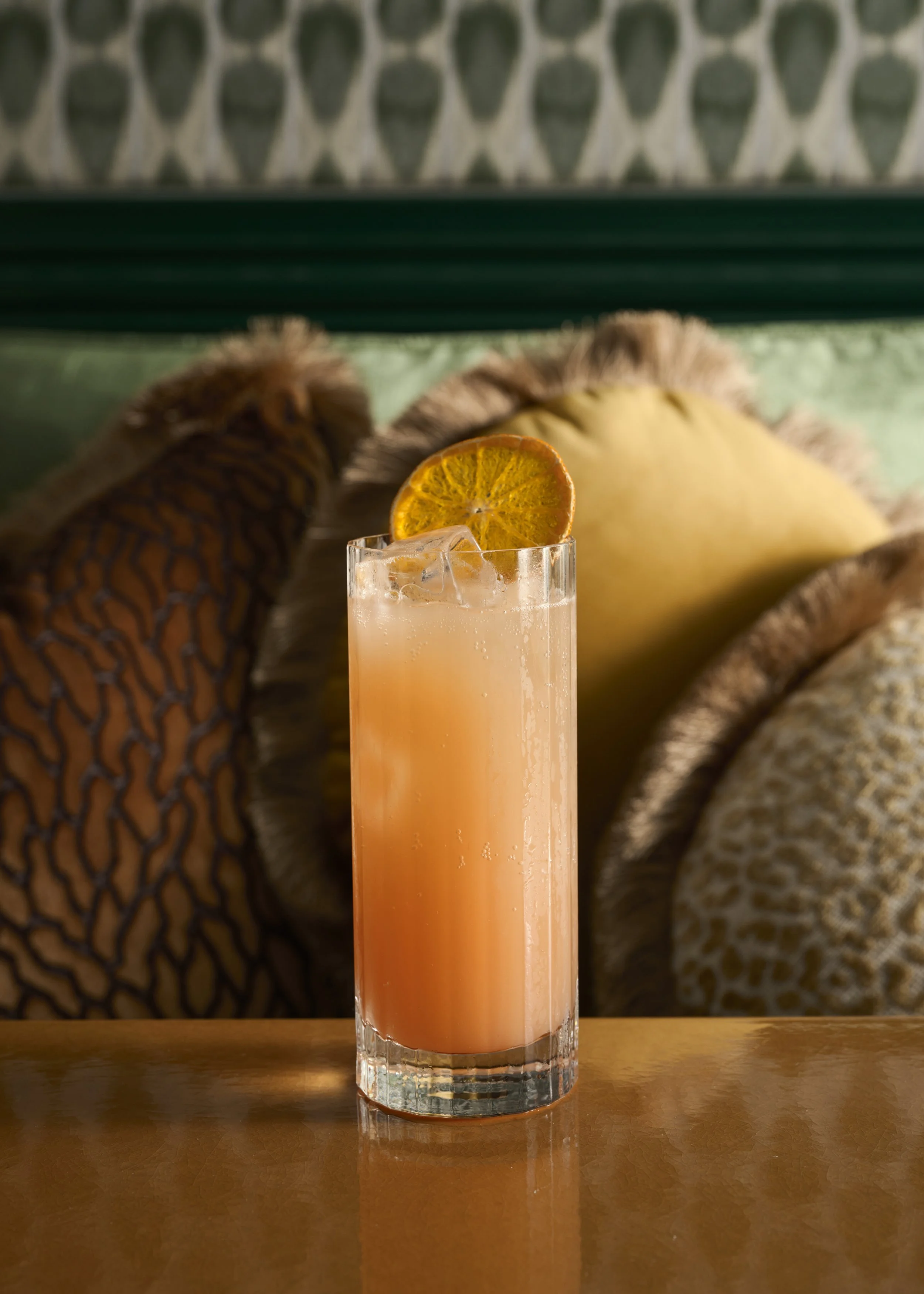 A tall glass of pink grapefruit juice with ice and a slice of dried orange on the rim, placed on a wooden surface.