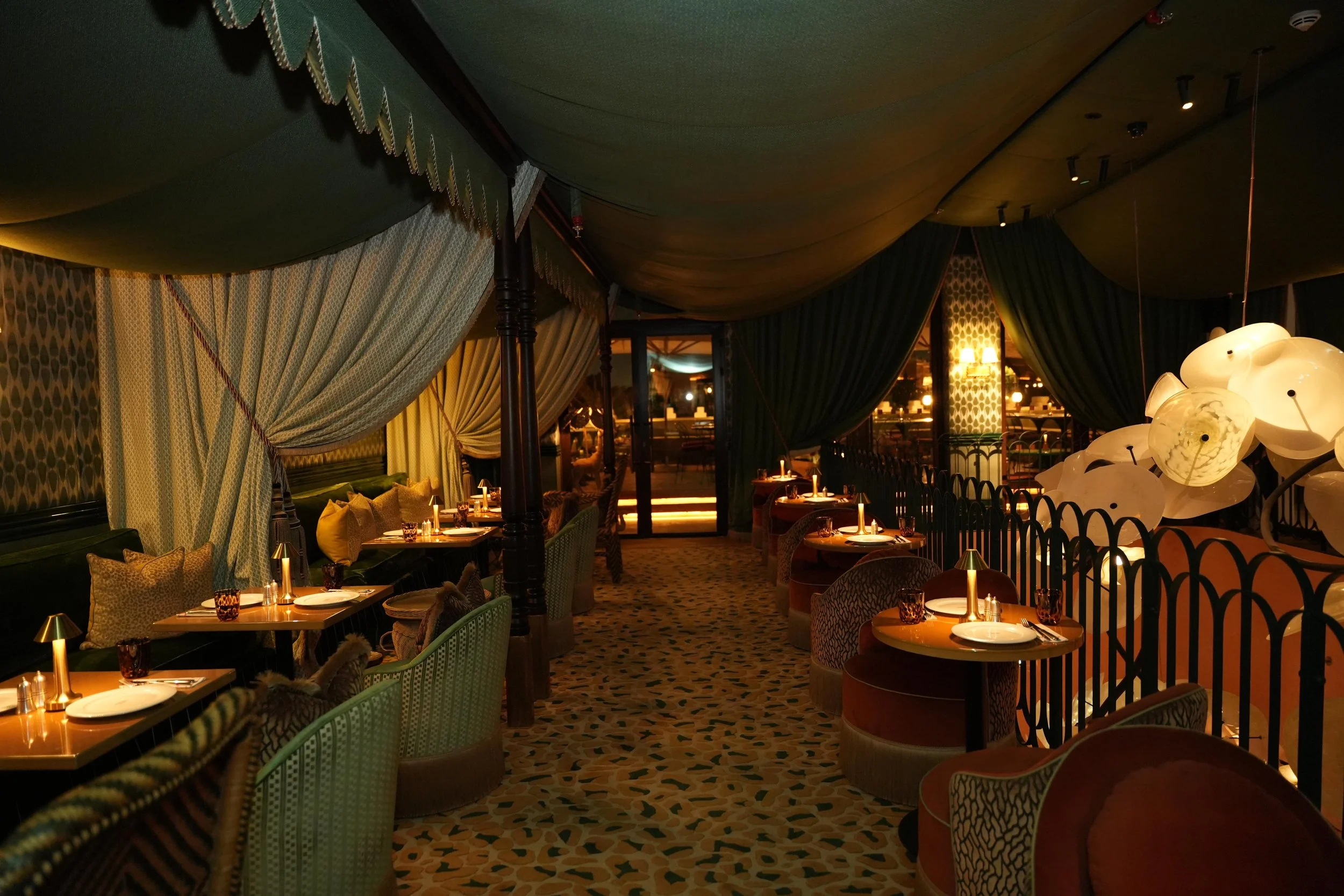A dimly lit restaurant with tables set for dining, surrounded by green and patterned fabric curtains and chairs, with soft lighting and decorative hanging lights.