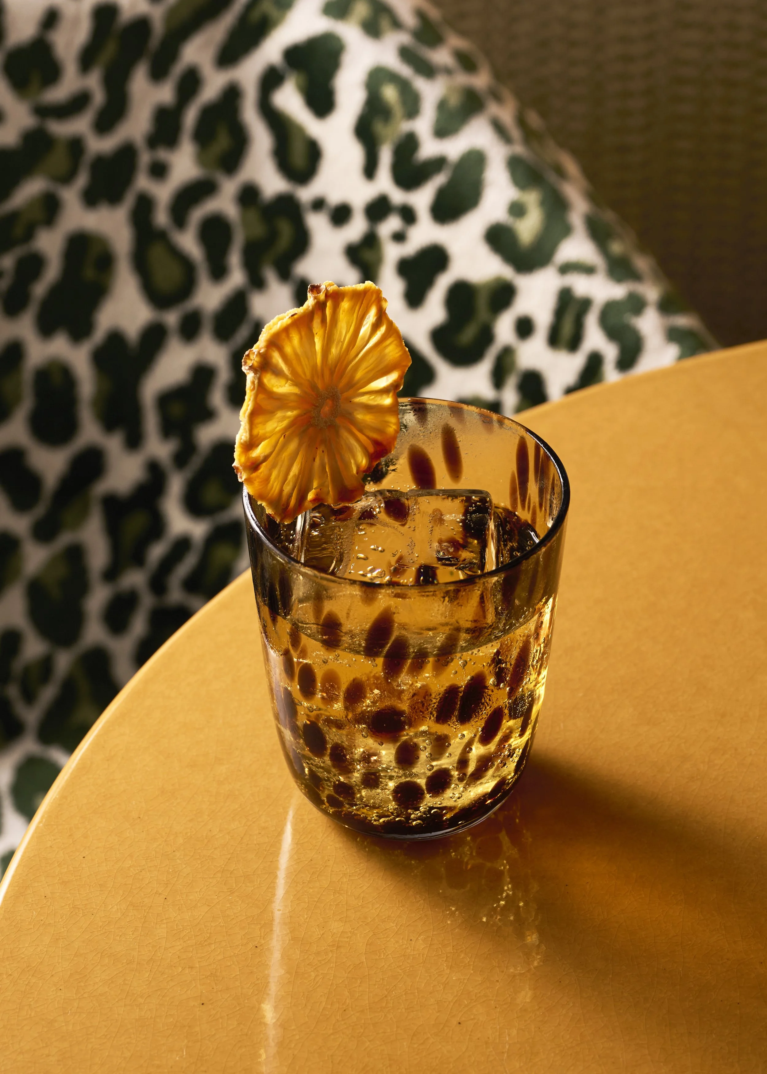 A glass of iced beverage with tapioca pearls, garnished with a dried pineapple slice, placed on a yellow table.