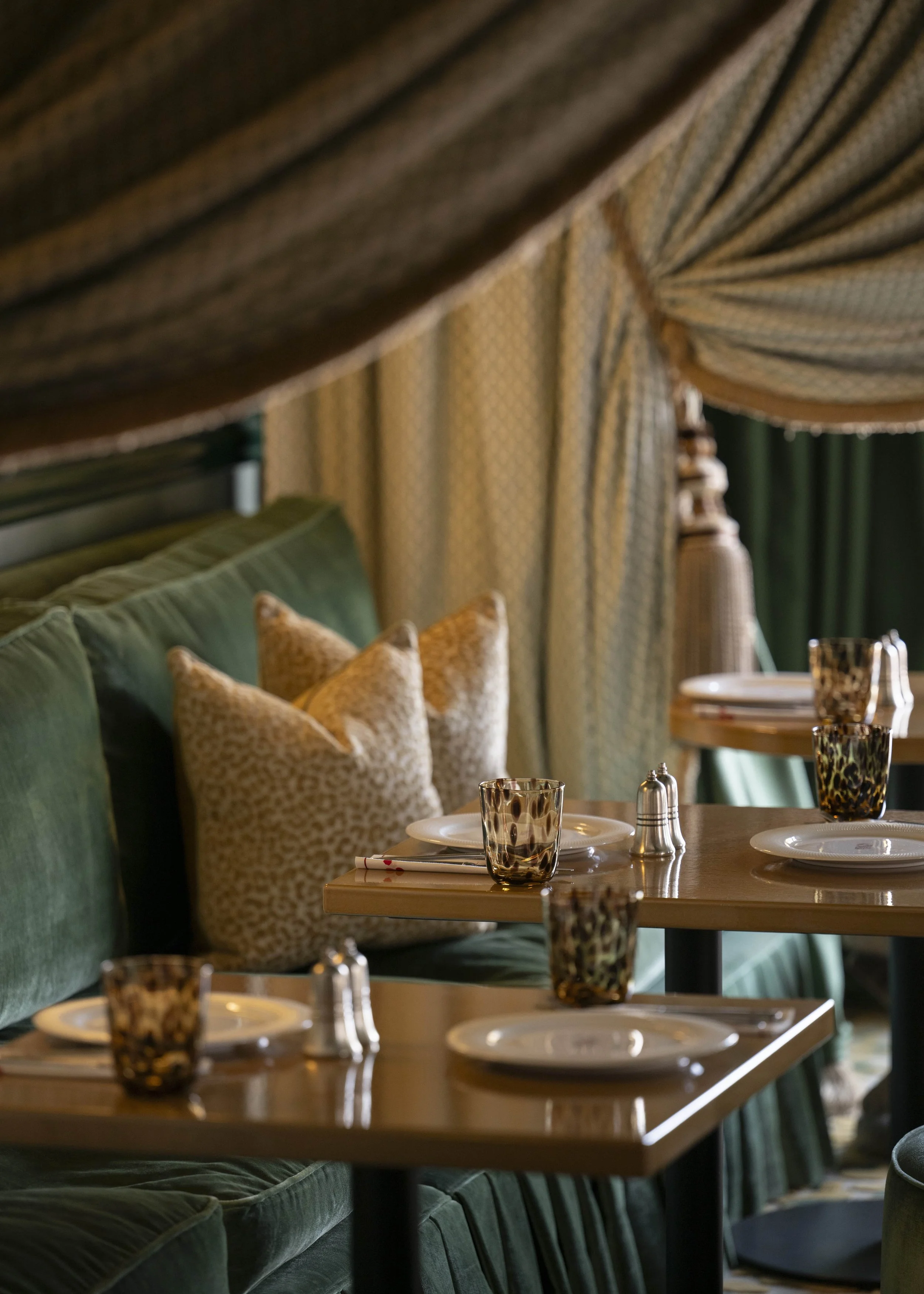 Elegant restaurant table setup with white plates, leopard print glassware, salt and pepper shakers, green cushioned seating, and patterned curtains in a warmly lit dining area.