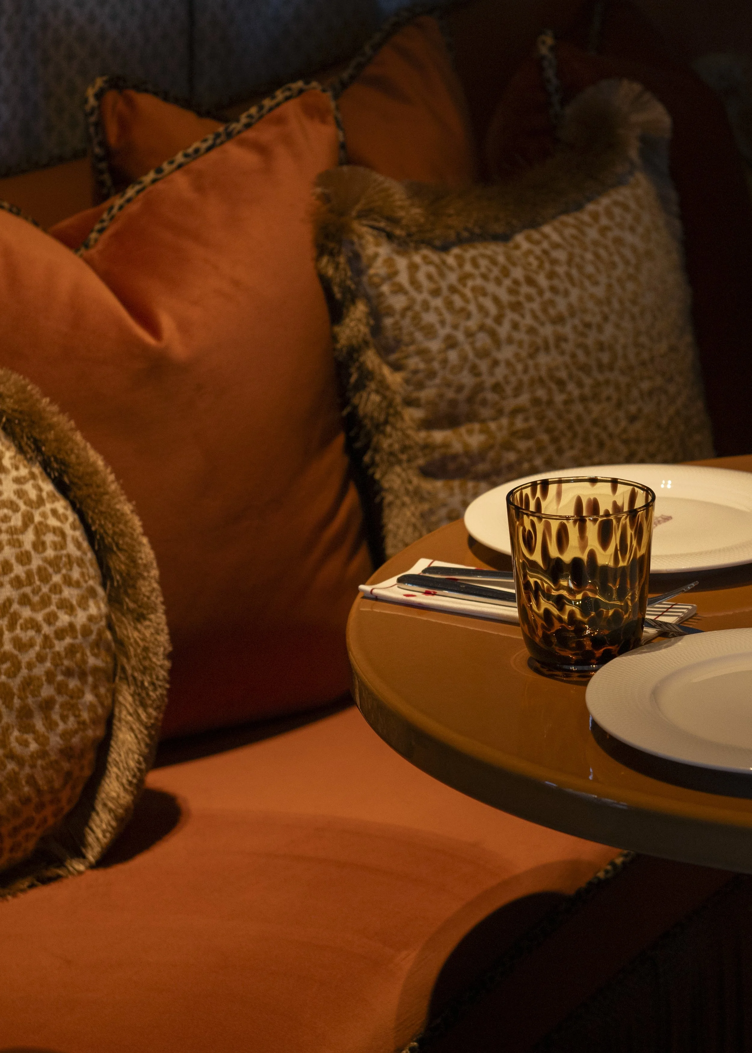 A cozy dining scene featuring a round wooden table set with white plates, a black-patterned glass, and silverware. Beige and leopard print cushions are arranged on a dark sofa in the background.