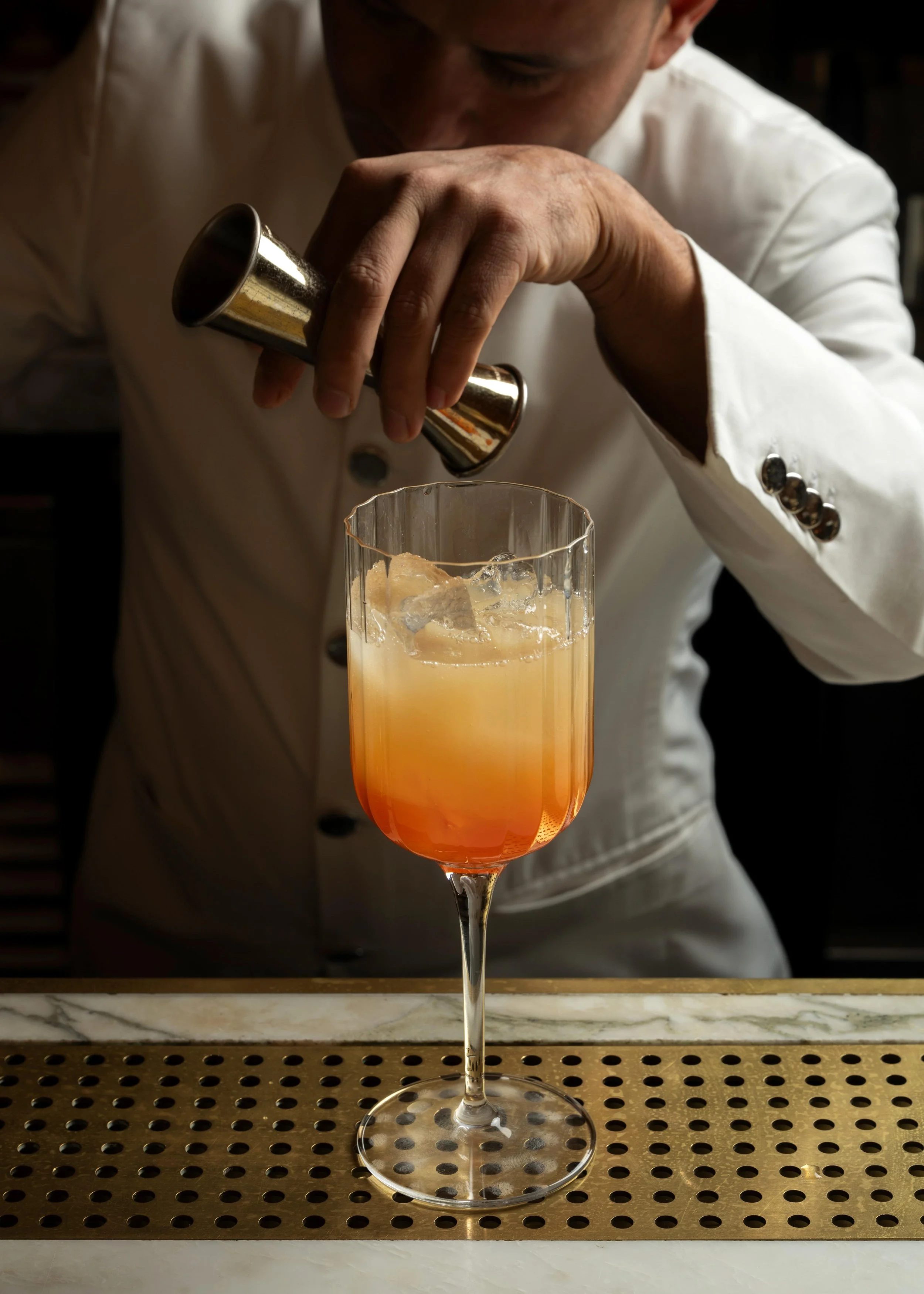 A bartender in a white jacket is adding a garnish or ingredient into a peach-colored cocktail with ice in a tall glass, on a bar counter.