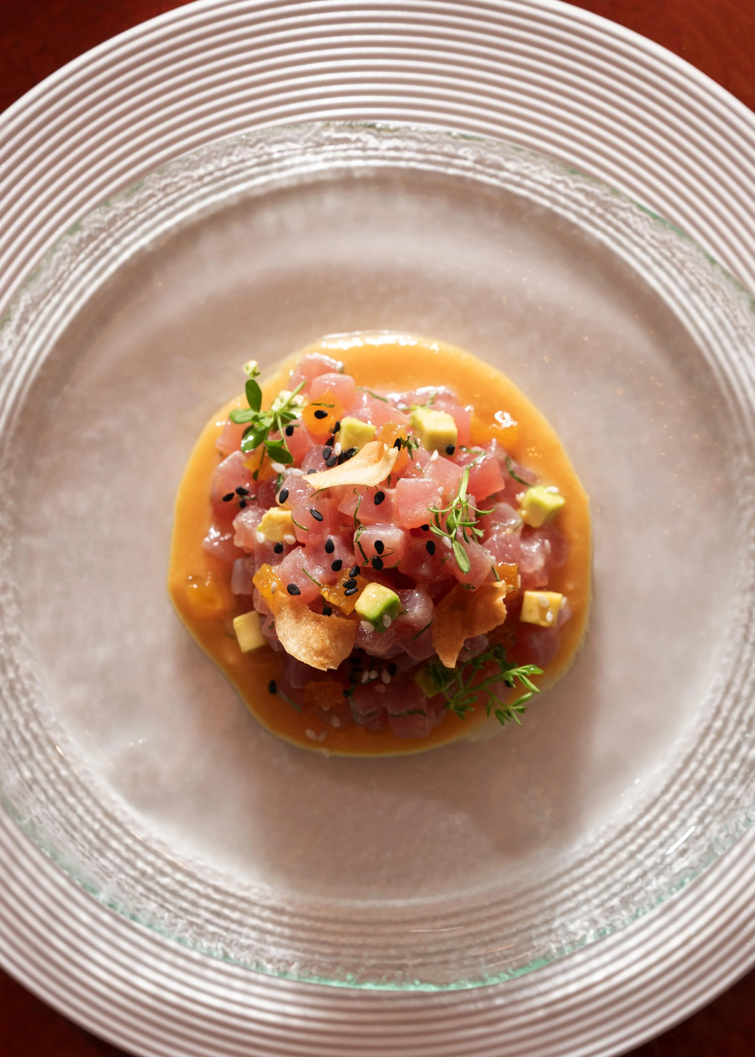 Tuna tartare garnished with microgreens, avocado, black sesame seeds, and crispy chips, served on a glass plate.