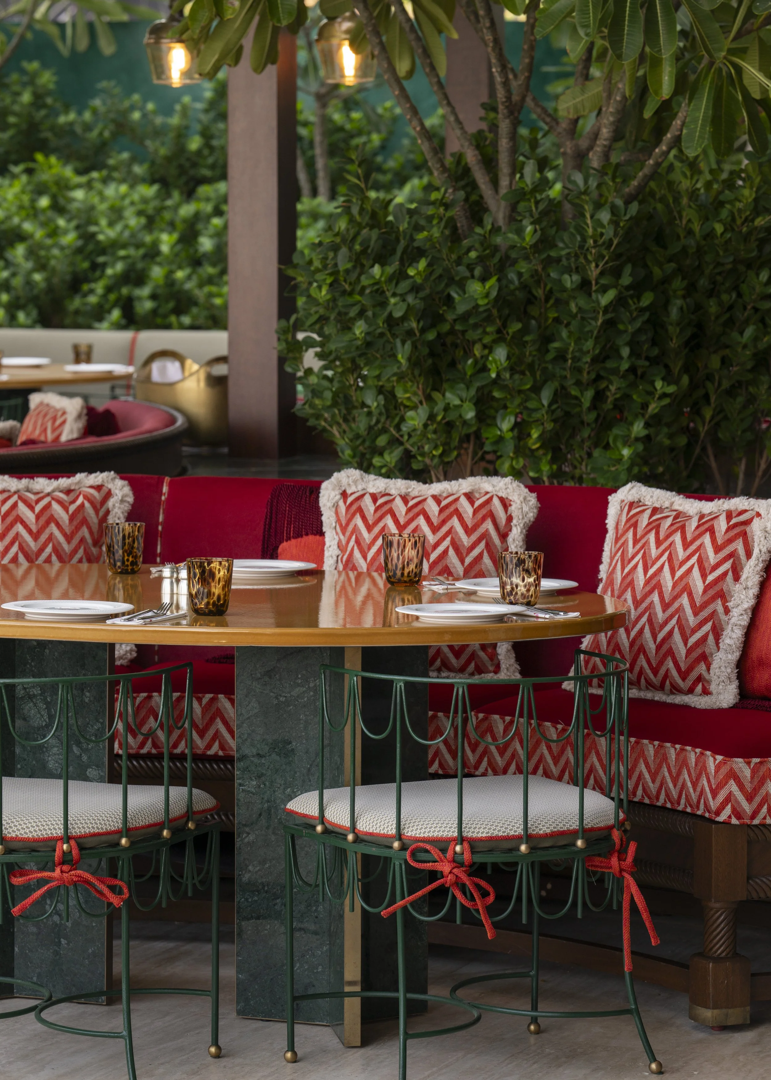 Outdoor dining area with a round wooden table, red upholstered seat, decorative patterned pillows, bronze for glassware, and green metal chairs with white cushions tied with red bows, lush green foliage in the background.
