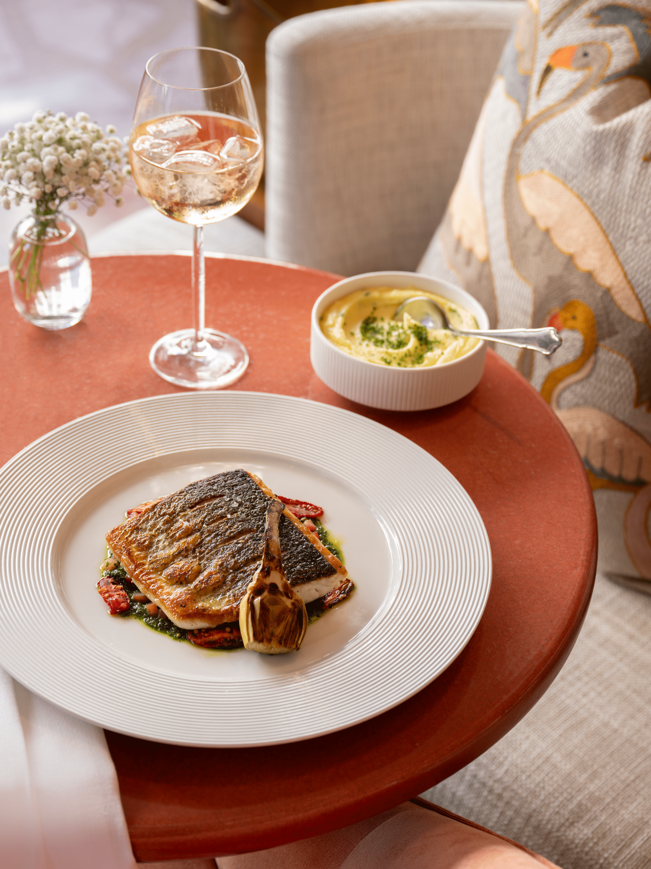 A dinner setup with a plate of cooked fish, a bowl of creamy soup, a glass of rosé wine, and a small vase with white flowers on a round, red-orange table.