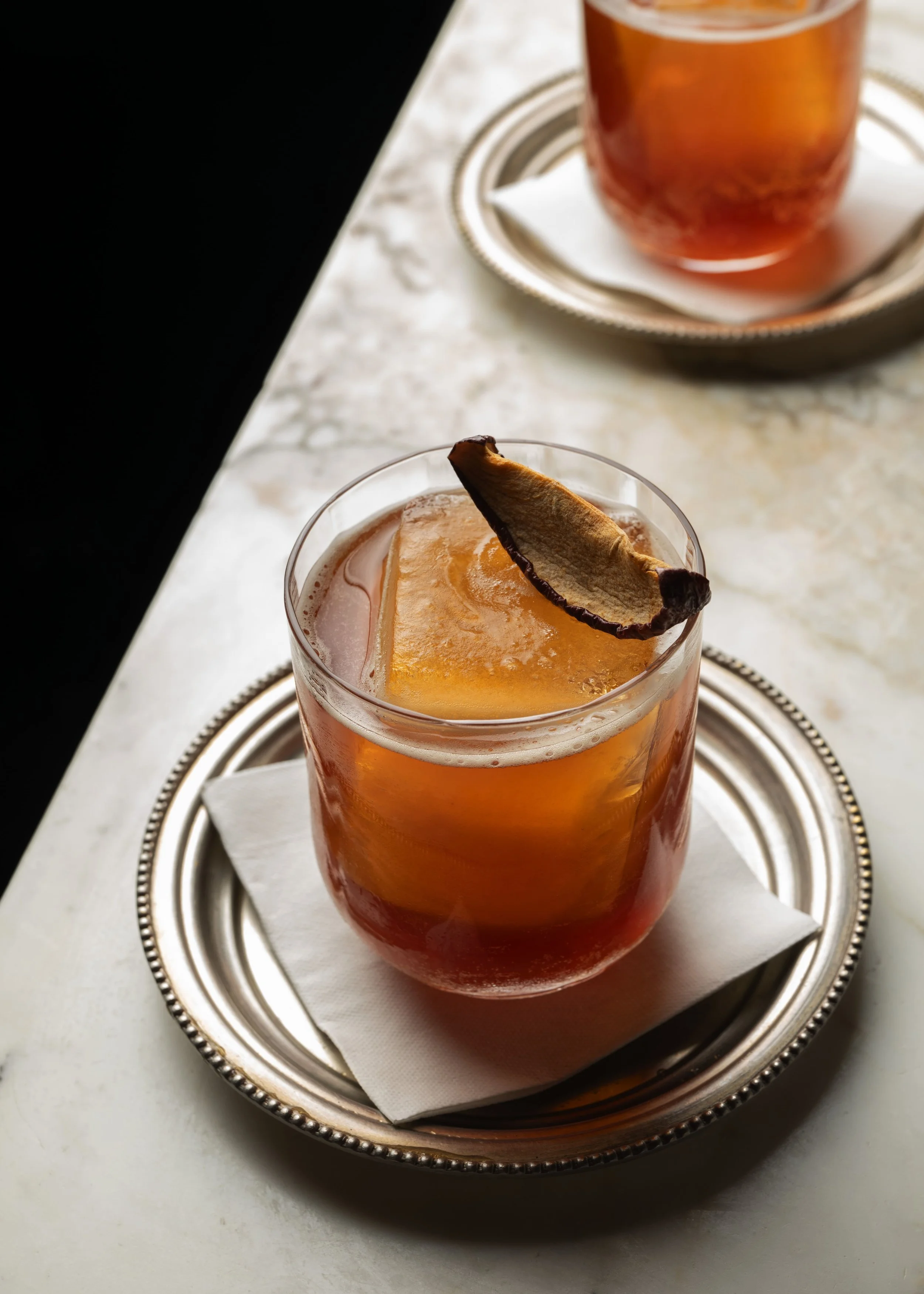 A glass of iced tea with a lemon slice and a dried lemon wheel garnish, placed on a silver tray with a napkin, on a white marble surface.