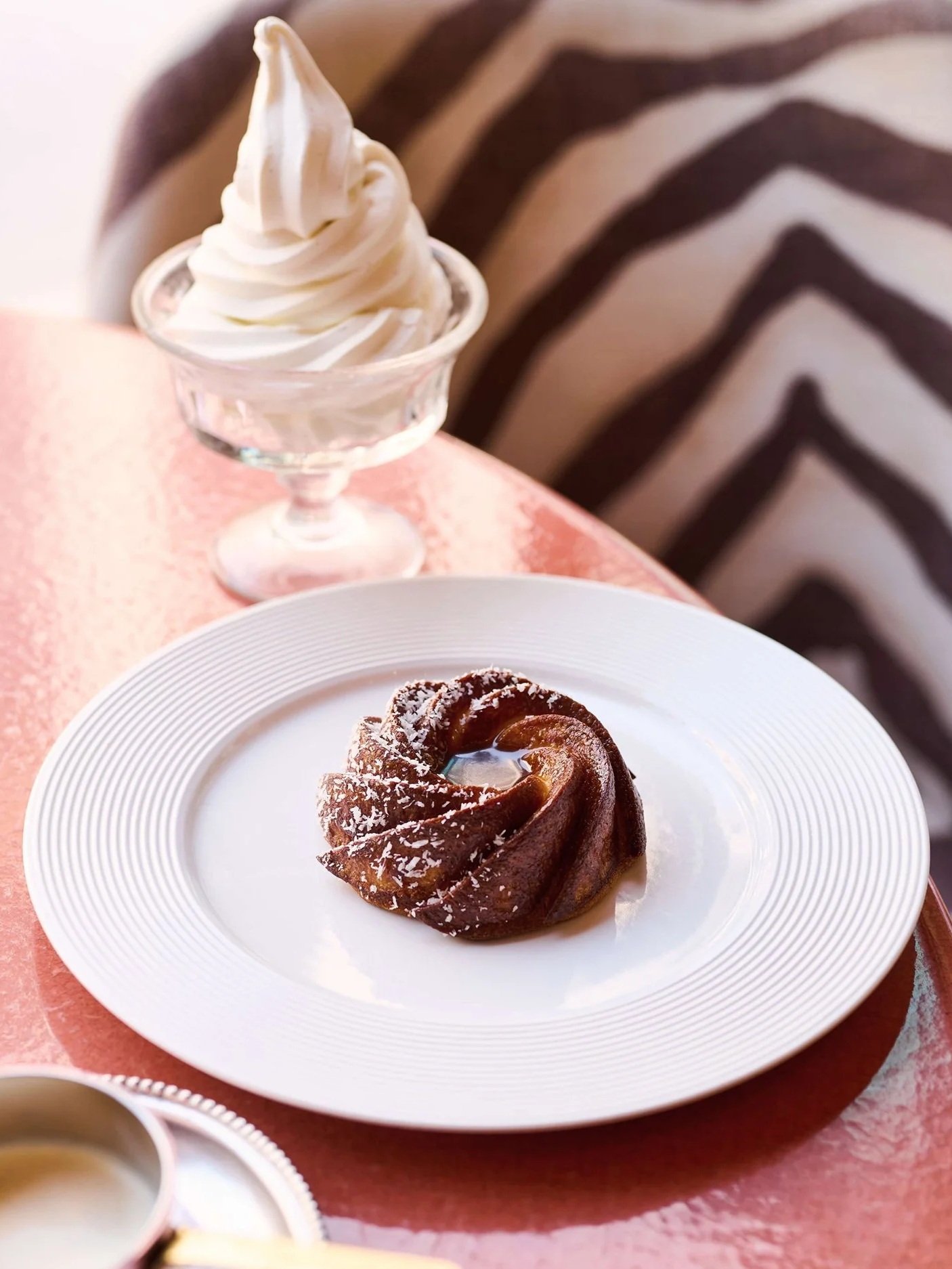 A serving of flan on a white plate and a glass of soft serve ice cream on a pink table.