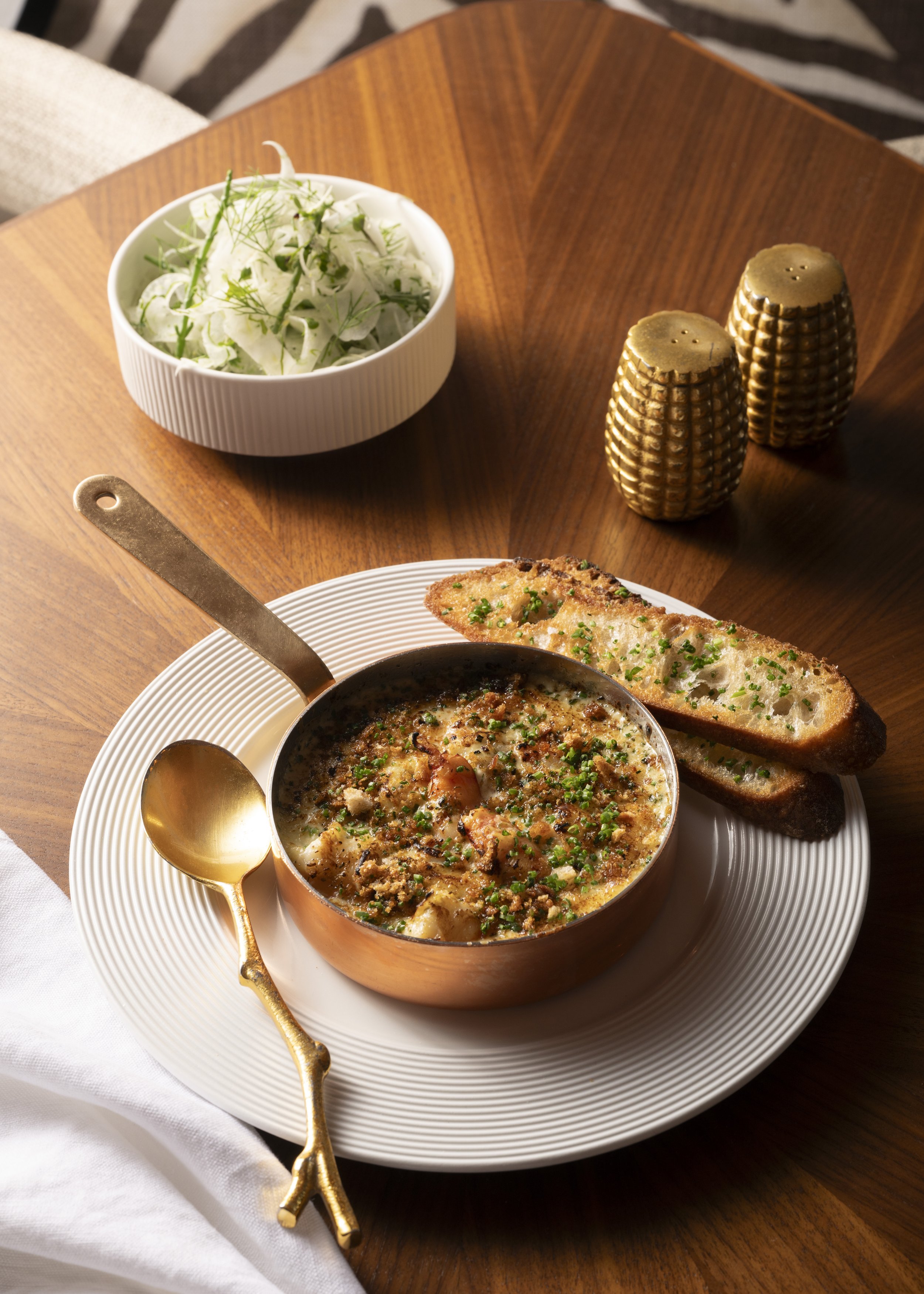 A meal of baked French onion soup garnished with chives, served with toasted bread slices, accompanied by a side salad with greens and herbs, on a wooden table with gold salt and pepper shakers.