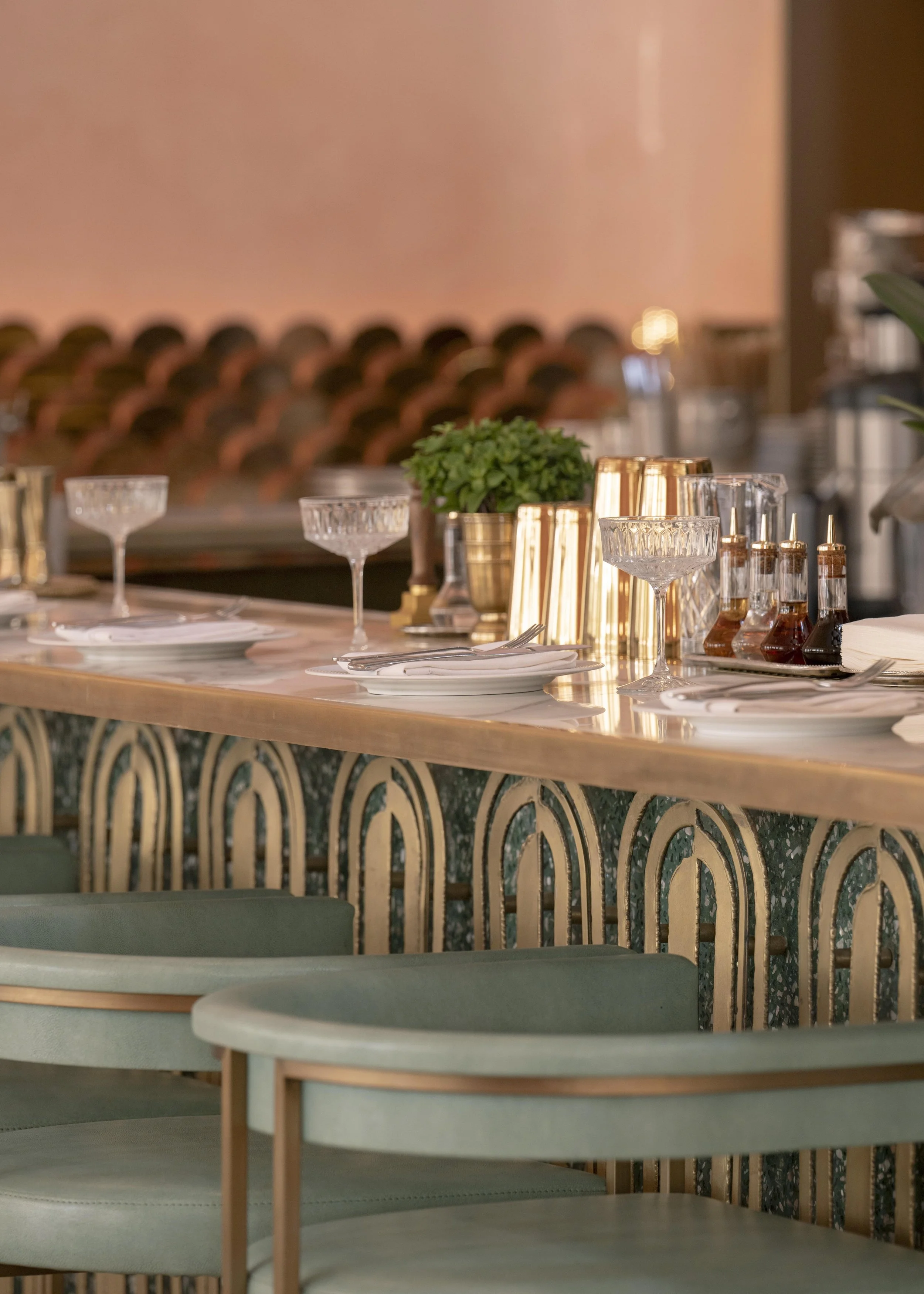 Elegant restaurant table setting with plates, glasses, cutlery, and condiments, with patterned decorative paneling on the counter and a potted plant, blurred dining area in the background.