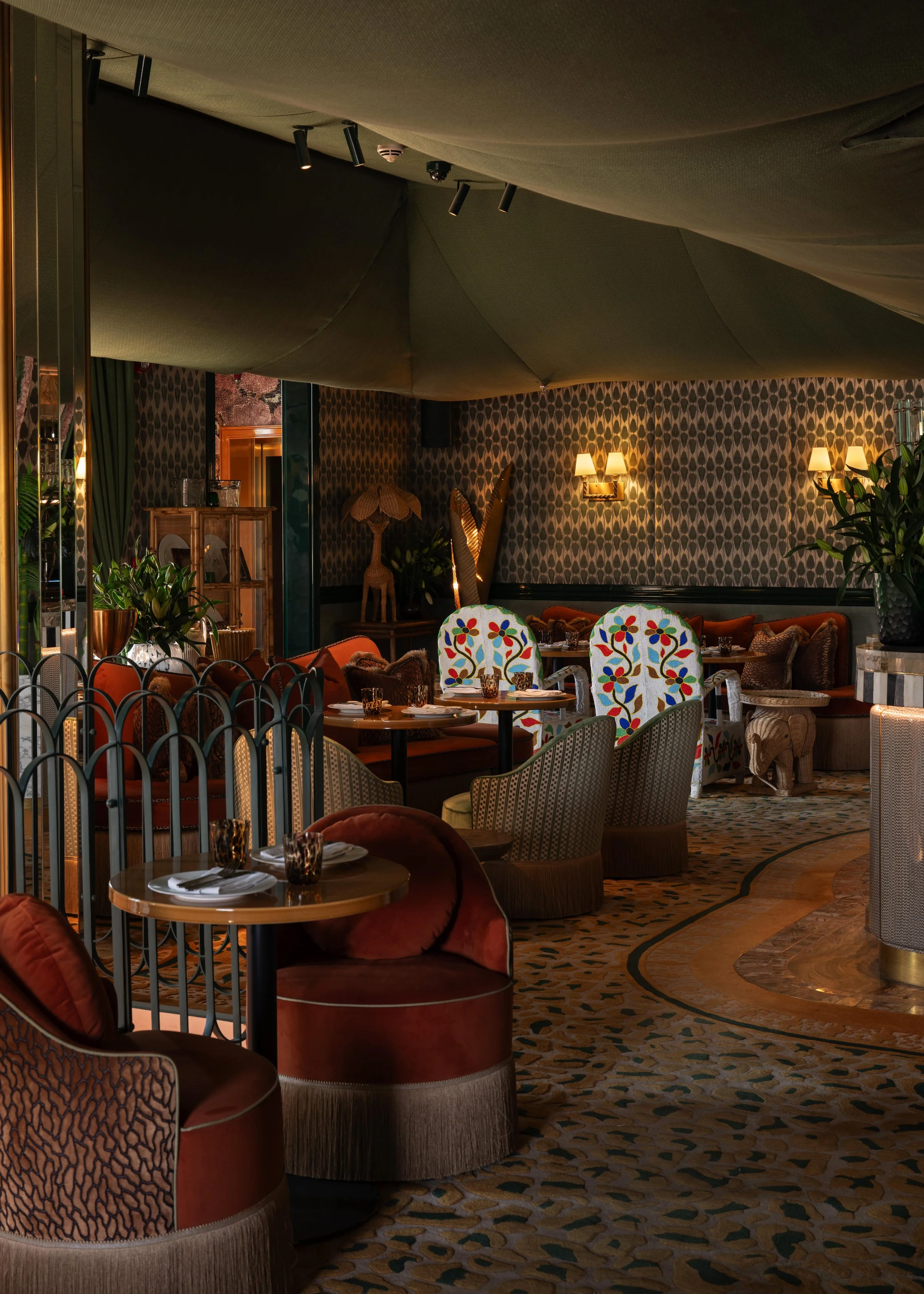 Interior view of a cozy restaurant or lounge with patterned wallpaper, various armchairs and sofas, small dining tables set with plates and glasses, decorative plants, warm lighting with wall sconces, and a textured carpet.