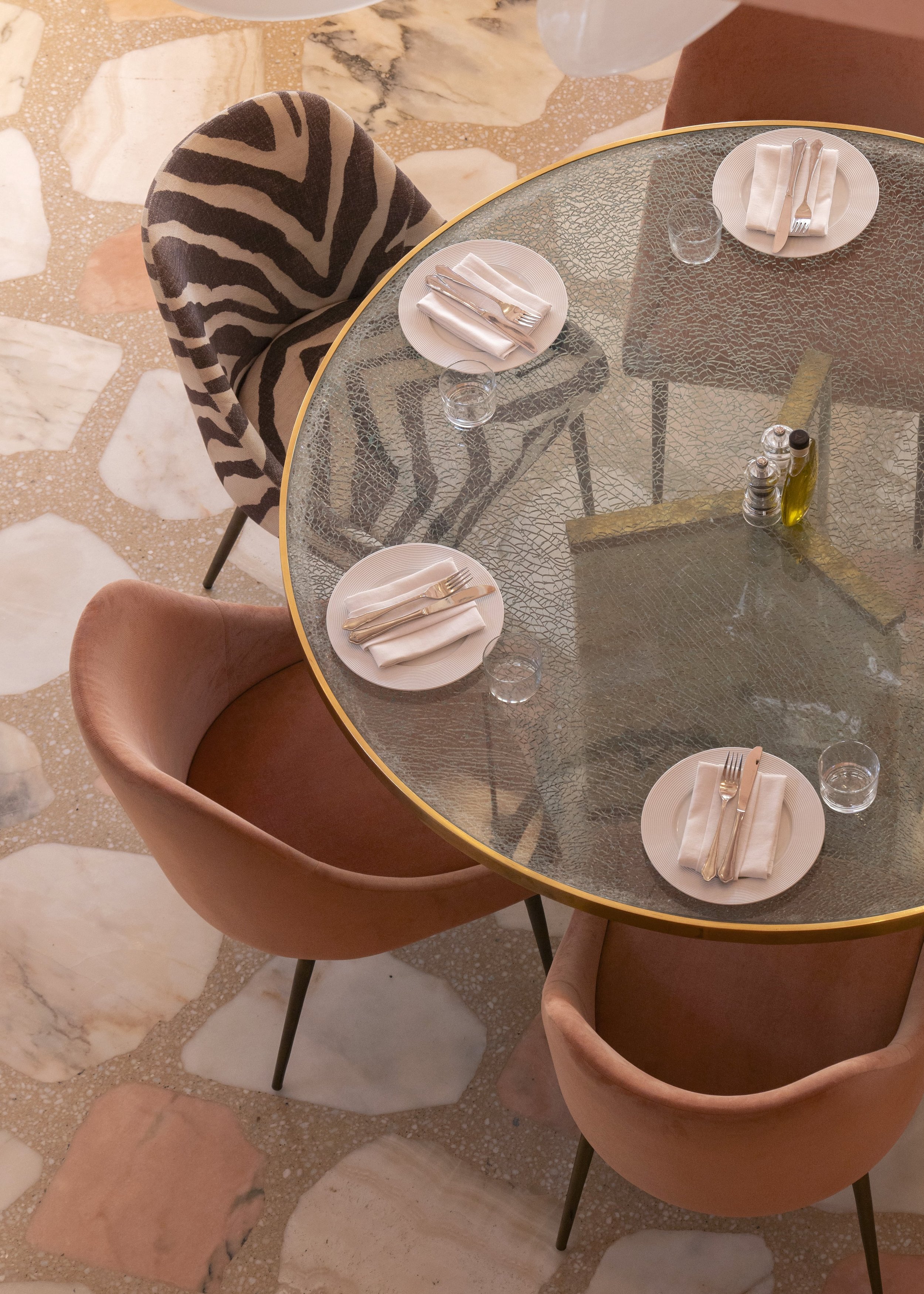 A round glass dining table set for four, with white plates, silverware, and small glasses. Four chairs surround the table: two pink velvet, one with a zebra print fabric, and the fourth one partially visible. There are also salt, pepper shakers, and 