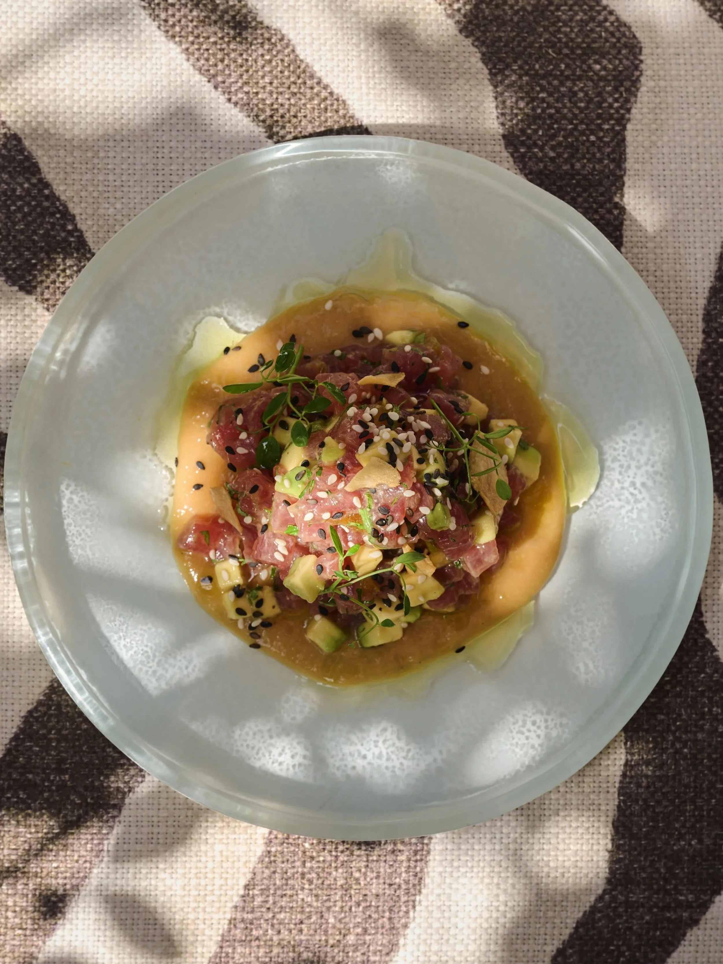 A plate of poke featuring diced raw fish, avocado, chopped green onions, microgreens, sesame seeds, and a sauce, served on a clear glass dish.
