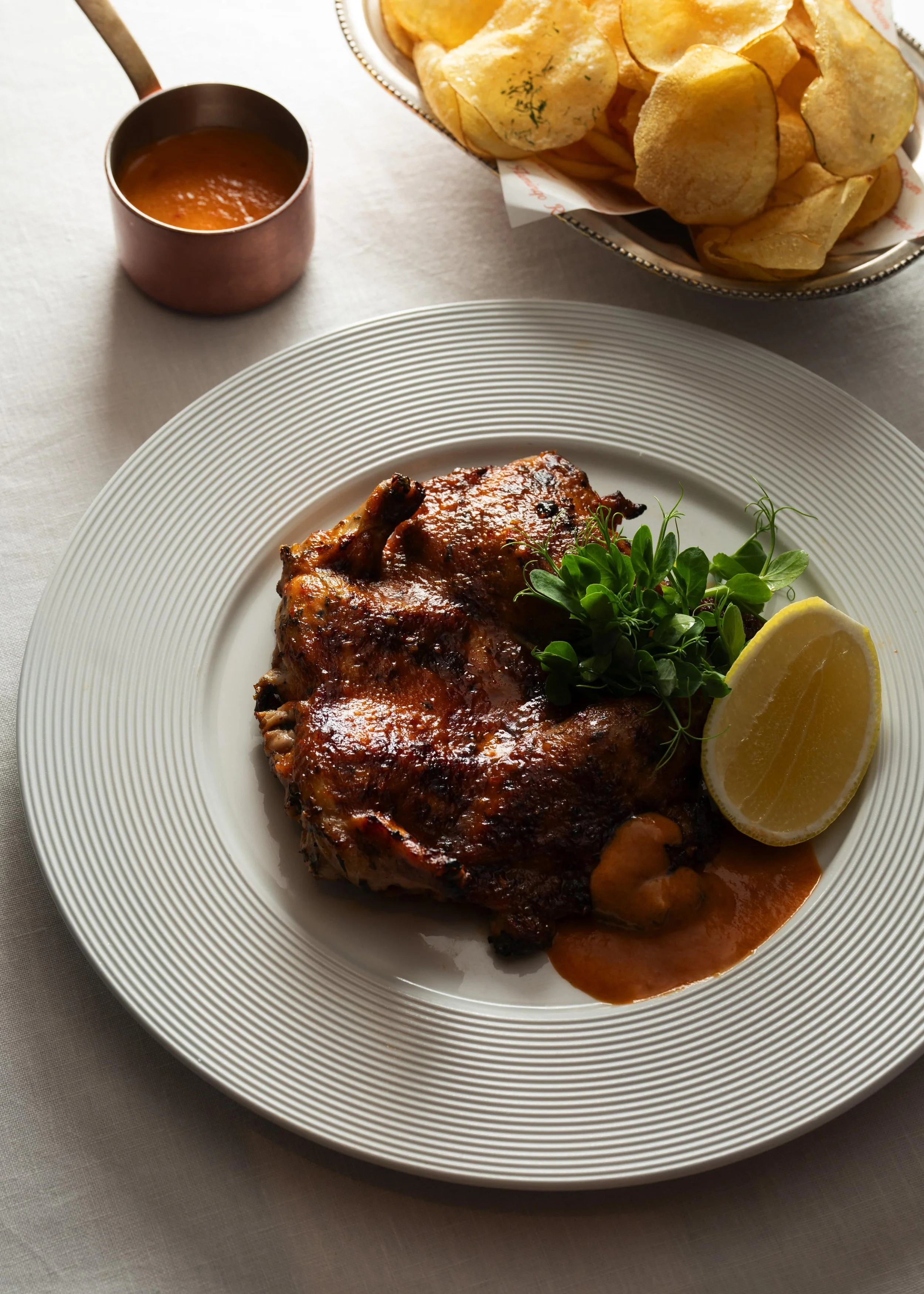 A plate of grilled chicken with barbecue sauce, a lemon wedge, and fresh herbs, with a side of potato chips and a cup of sauce in the background.