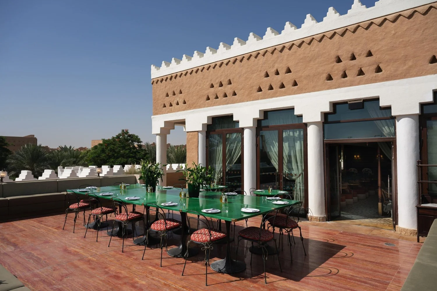 Outdoor dining area with a large green table set with plates, glasses, and cutlery, surrounded by chairs with red and black patterned cushions, on a terracotta-tiled patio next to a building with white columns and a Moroccan-style design.