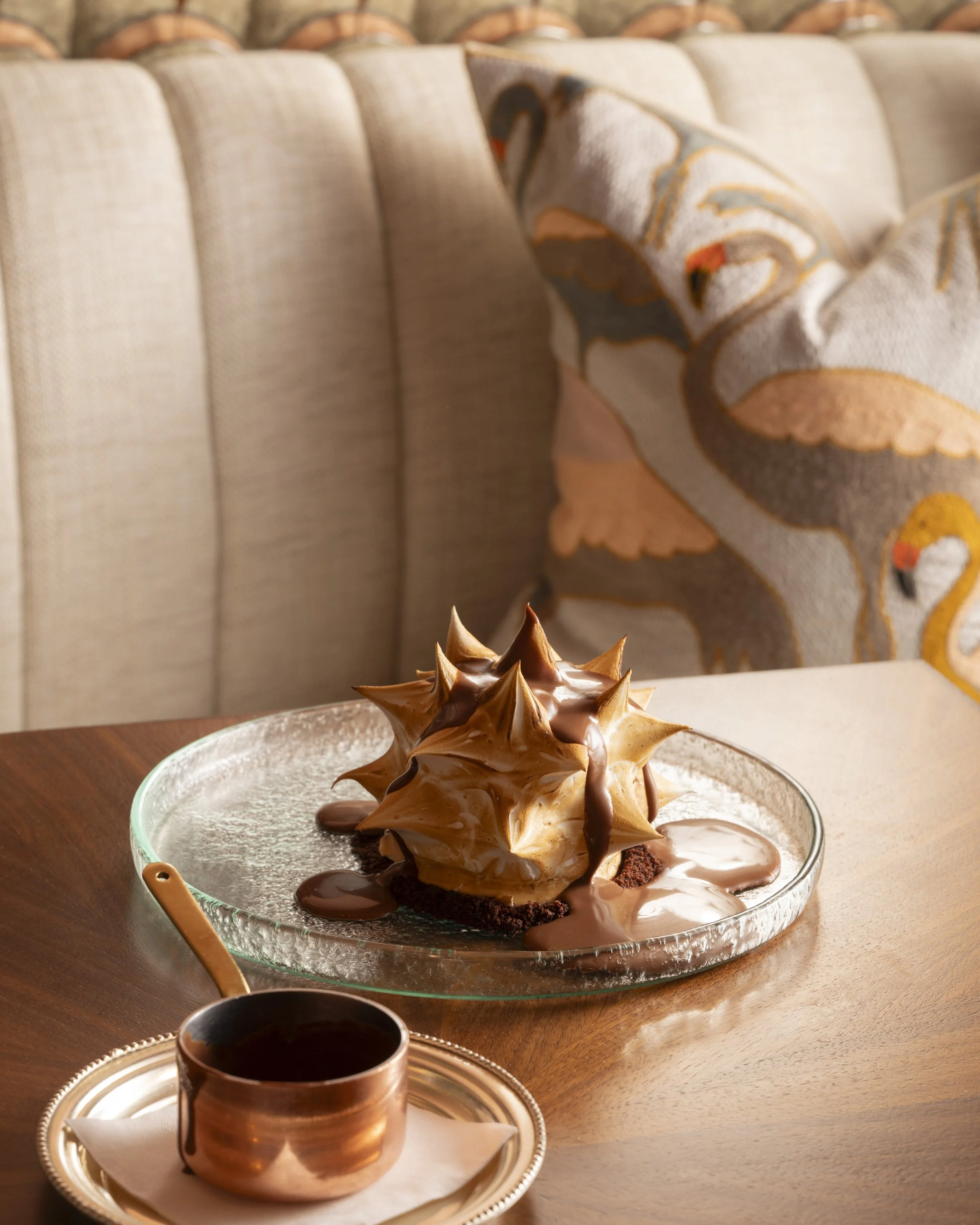 A glass plate with a large chocolate and toasted meringue dessert, accompanied by a cup of hot chocolate on a gold tray, set on a wooden table with a decorative pillow with swan and floral patterns in the background.