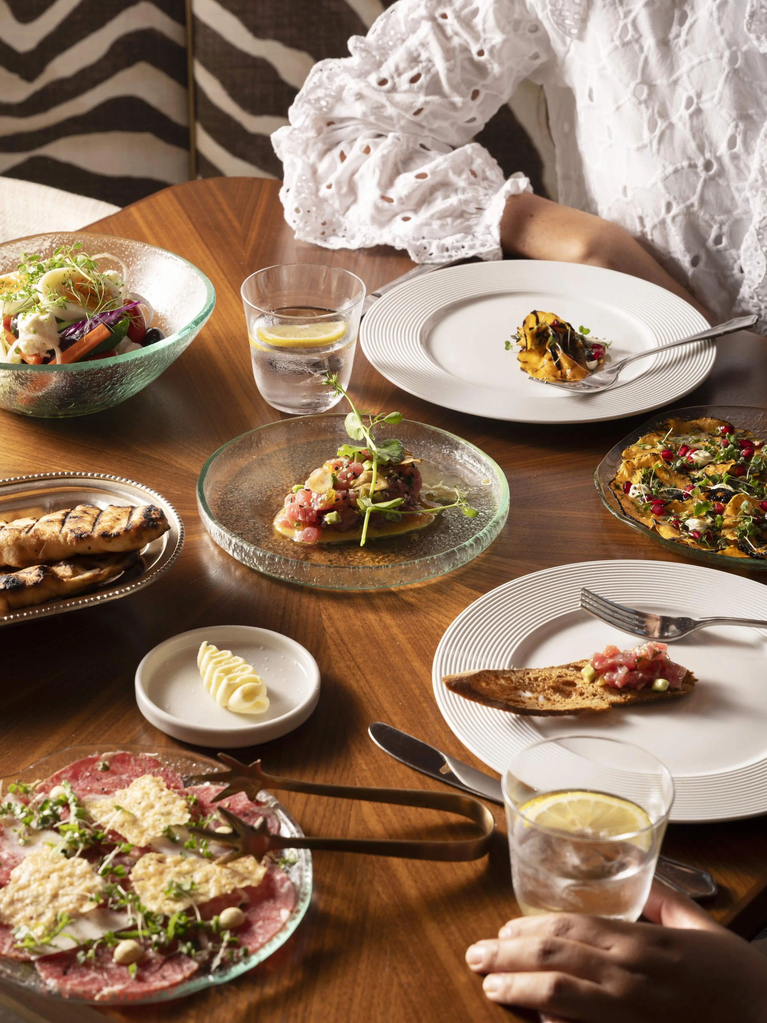 A dining table set with various dishes, including a salad, grilled meats, a tart, and bread, with glasses of water and a hand holding a lemon wedge.