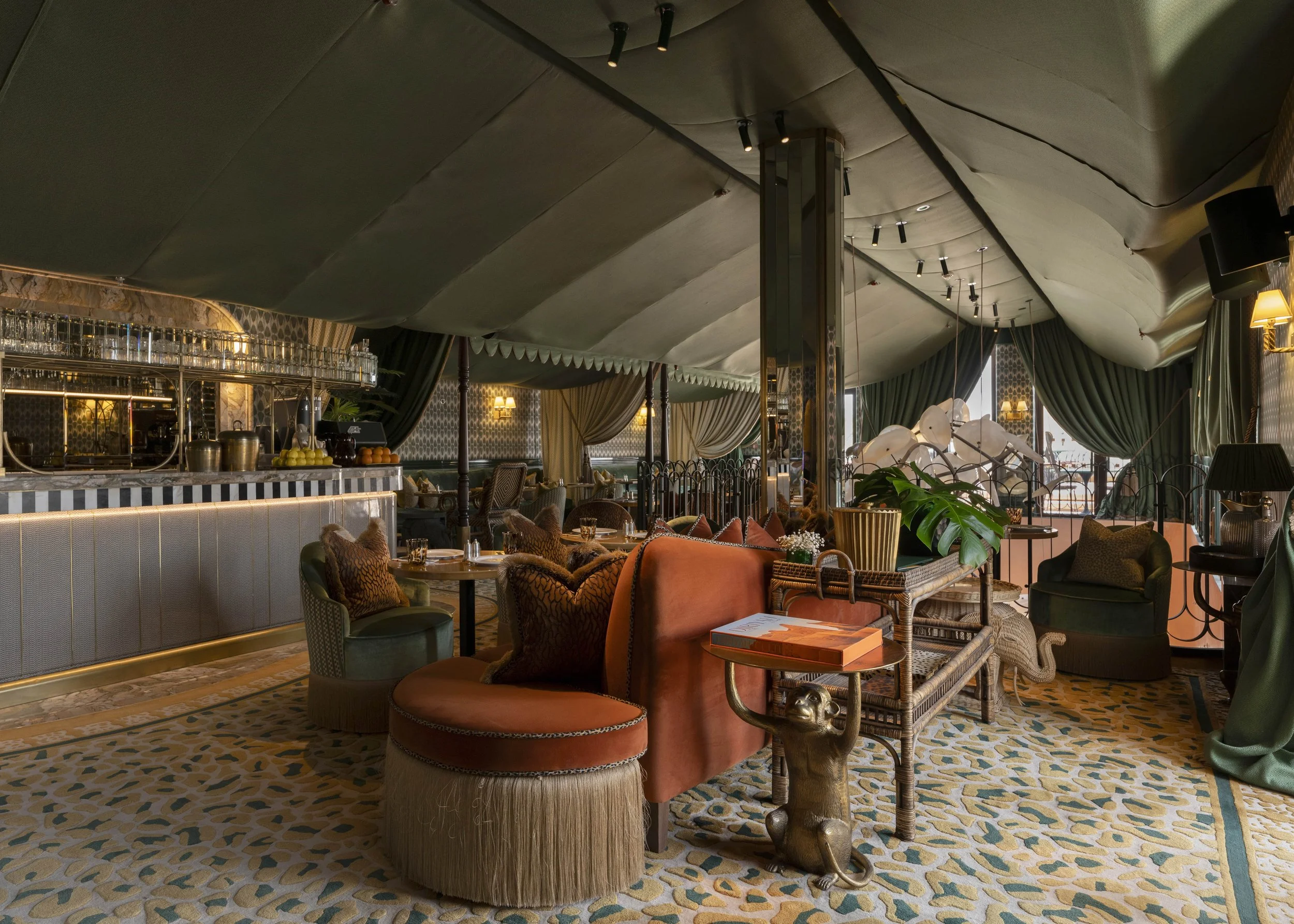 Luxurious restaurant interior with green drapes, patterned carpet, and eclectic furniture including a pink velvet chair, a green chair, and a wicker bench with a gold elephant sculpture at its side. The space is decorated with plants, books, and ambi