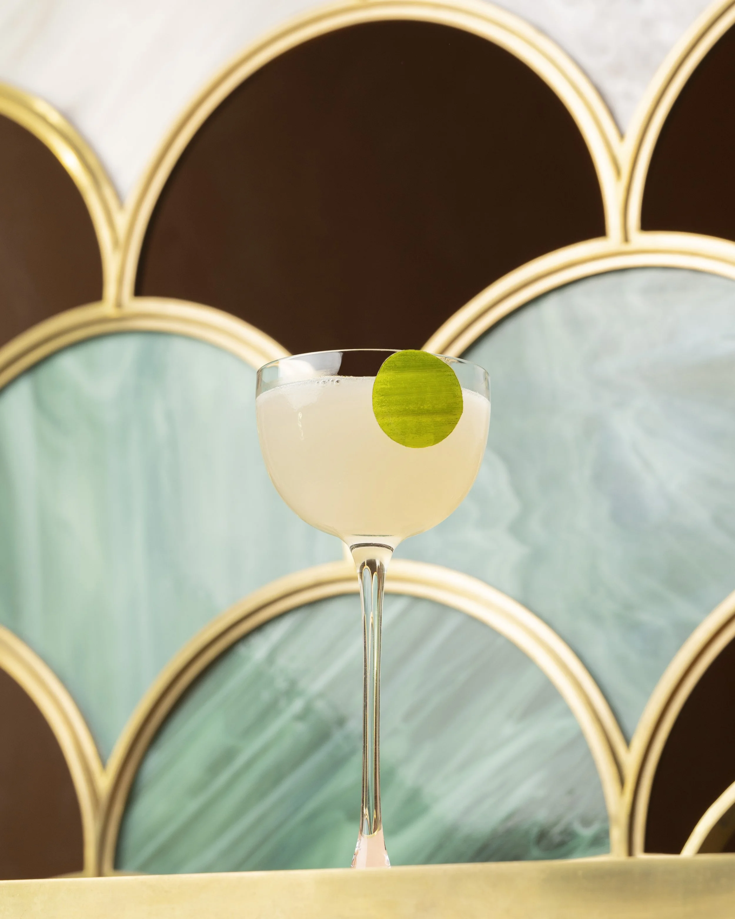 A cocktail glass with a pale yellow drink and a lime wedge garnish, set against a decorative background of overlapping curved panels with gold trim.