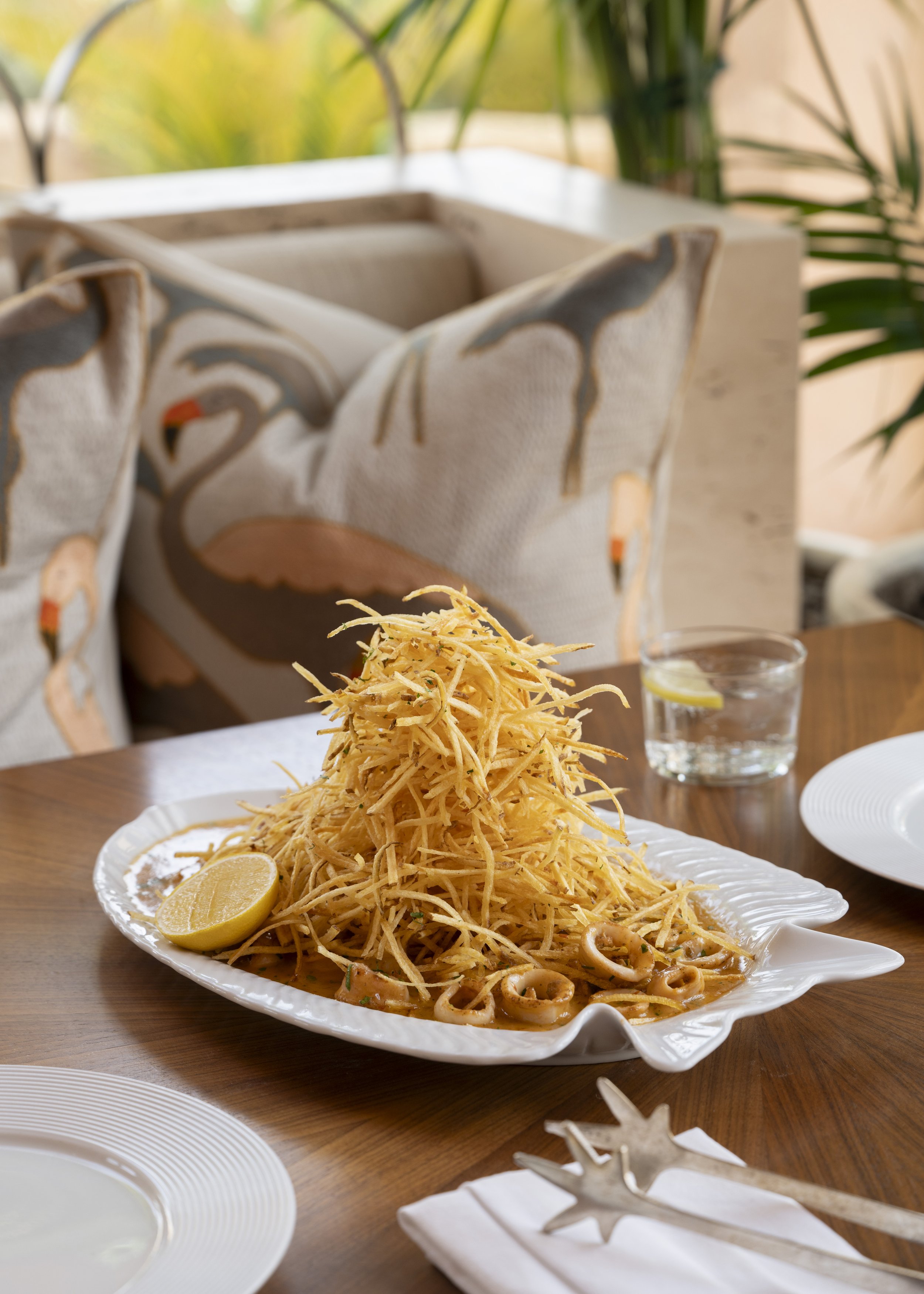 Plate of fried food, possibly fish and chips or shredded potatoes, garnished with lemon wedges, on a dining table with glass of water and white plates, in a bright setting with tropical decor.