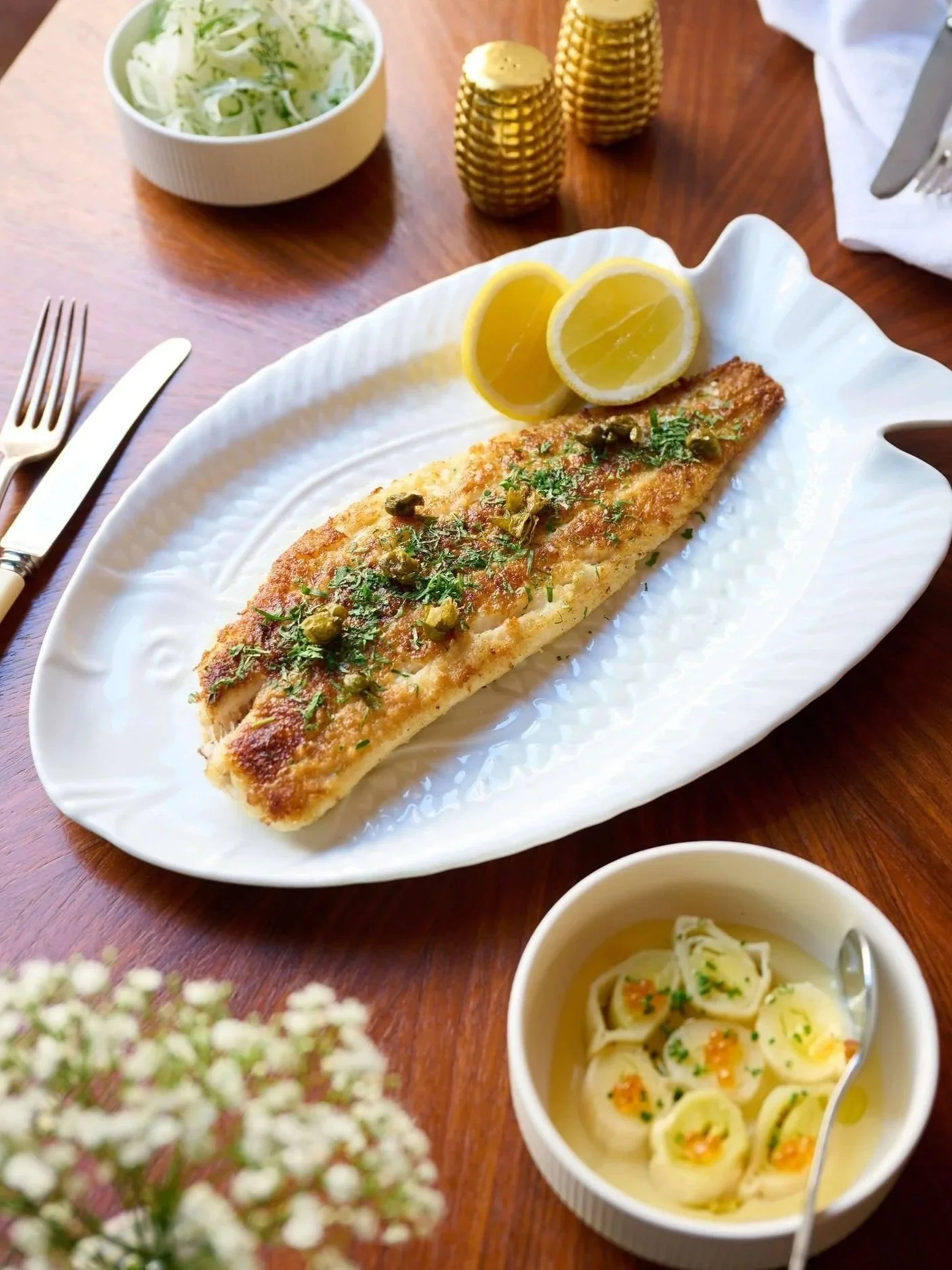 A plated serving of grilled fish garnished with herbs and capers, with lemon slices on the side, on a white decorative plate. There is a bowl of salad with shredded lettuce, a small bowl of tortellini in broth, and a set of gold salt and pepper shakers on a wooden table.