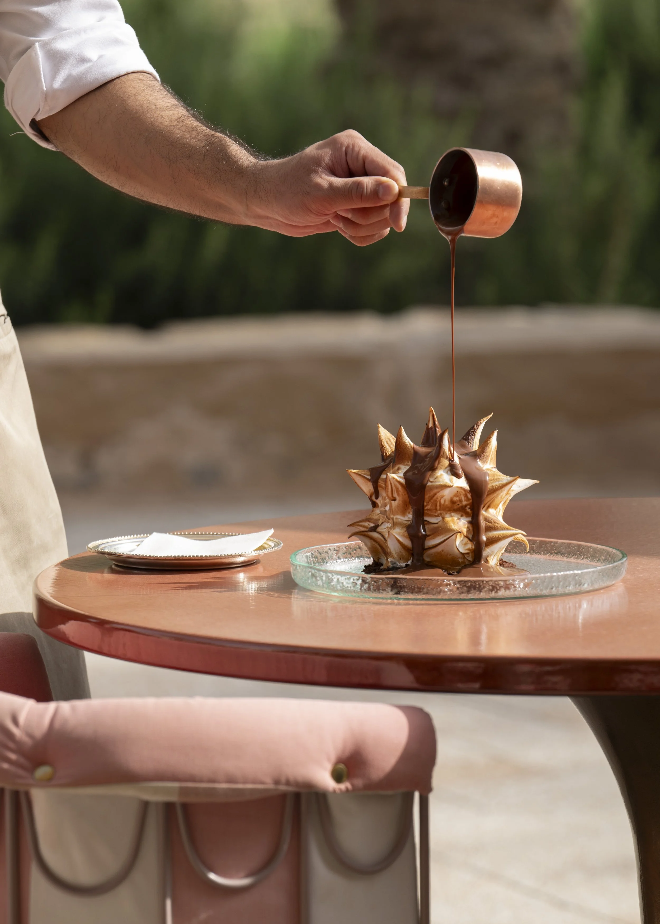 A person is pouring chocolate sauce over a meringue dessert on a glass plate, with the setting outdoors near greenery.