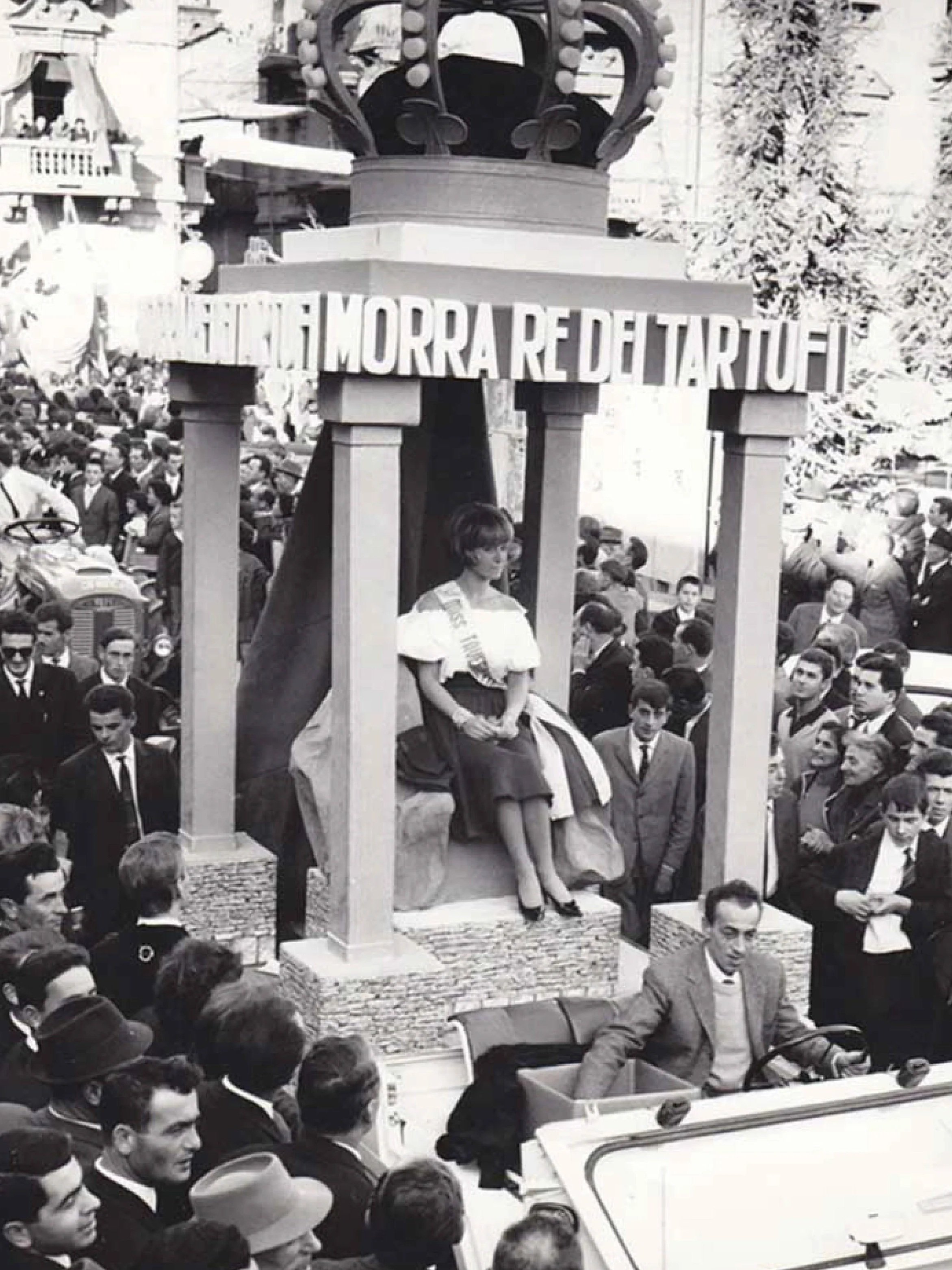 Black and white photo of a parade with a woman seated on a decorated float labeled "MORRA RE DEL TARTUFI," surrounded by a crowd of men and women in formal attire.