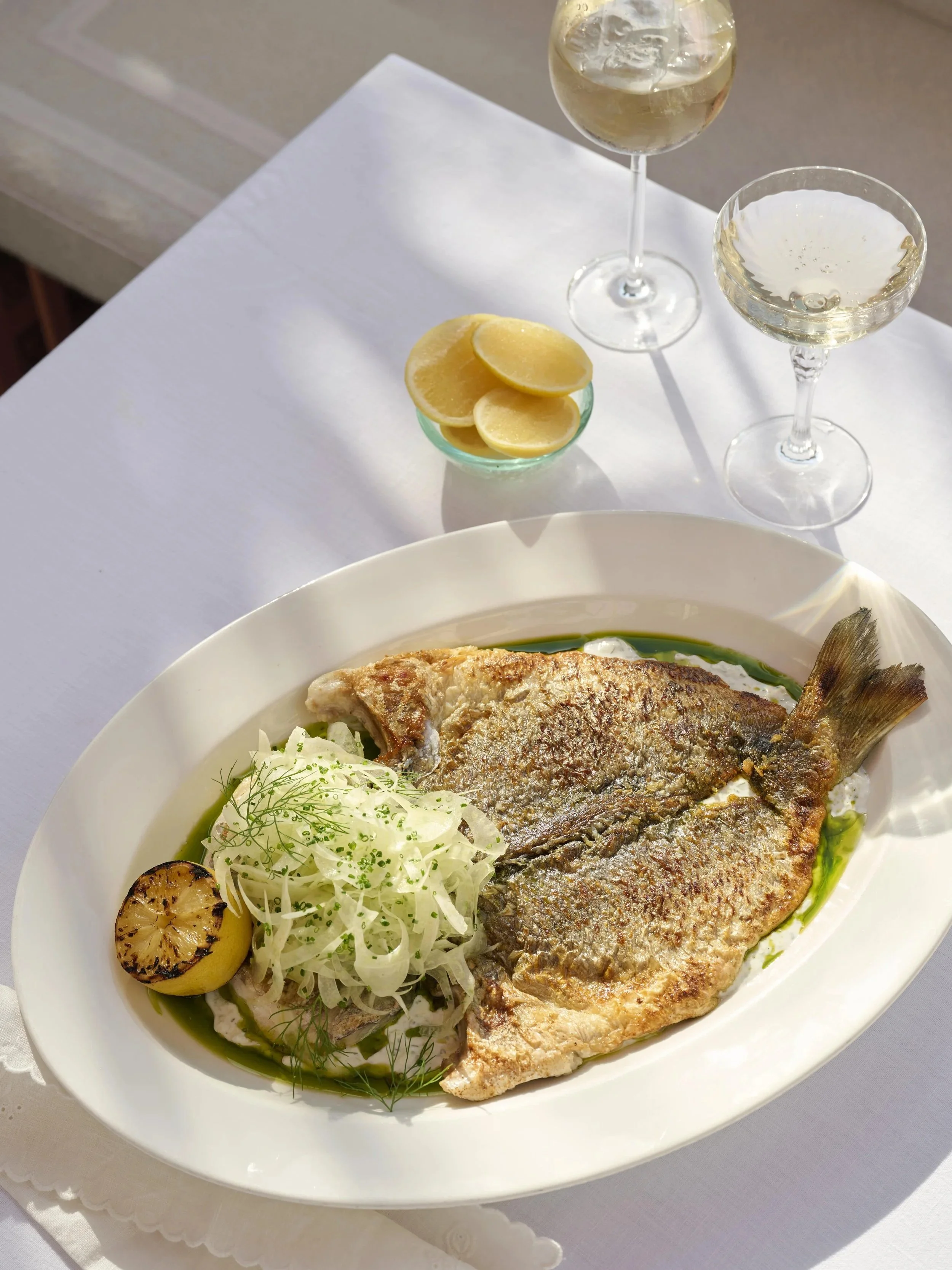 A whole fried fish on a white oval plate, garnished with sliced fennel and herbs, with a lemon half and a grilled lemon wedge on the side, placed on a white tablecloth. In the background, there are two glasses of white wine, a glass of clear beverage, and a small bowl of lemon slices.