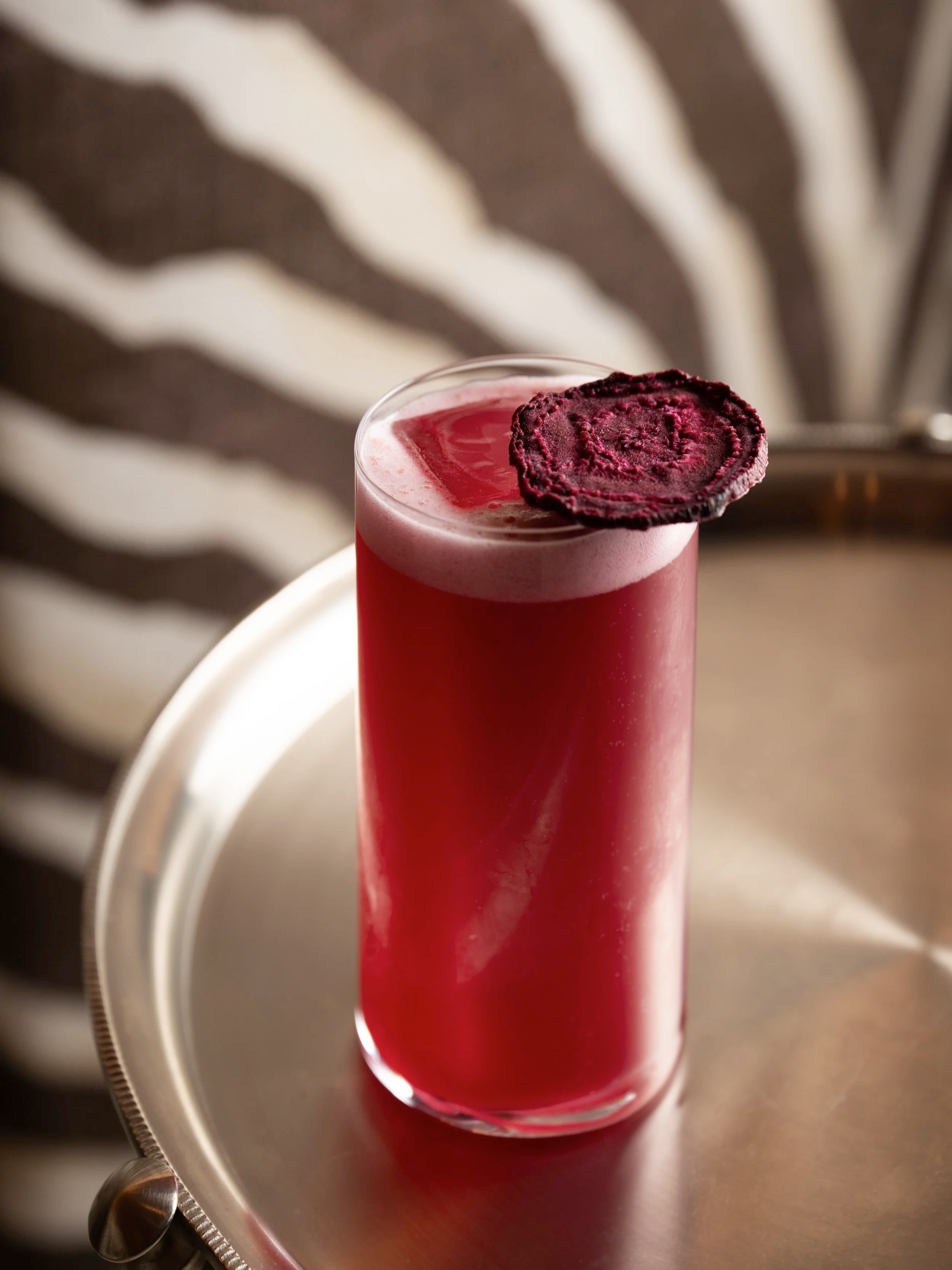 A tall glass of pink and purple layered cocktail topped with a purple edible flower garnish, placed on a silver tray.