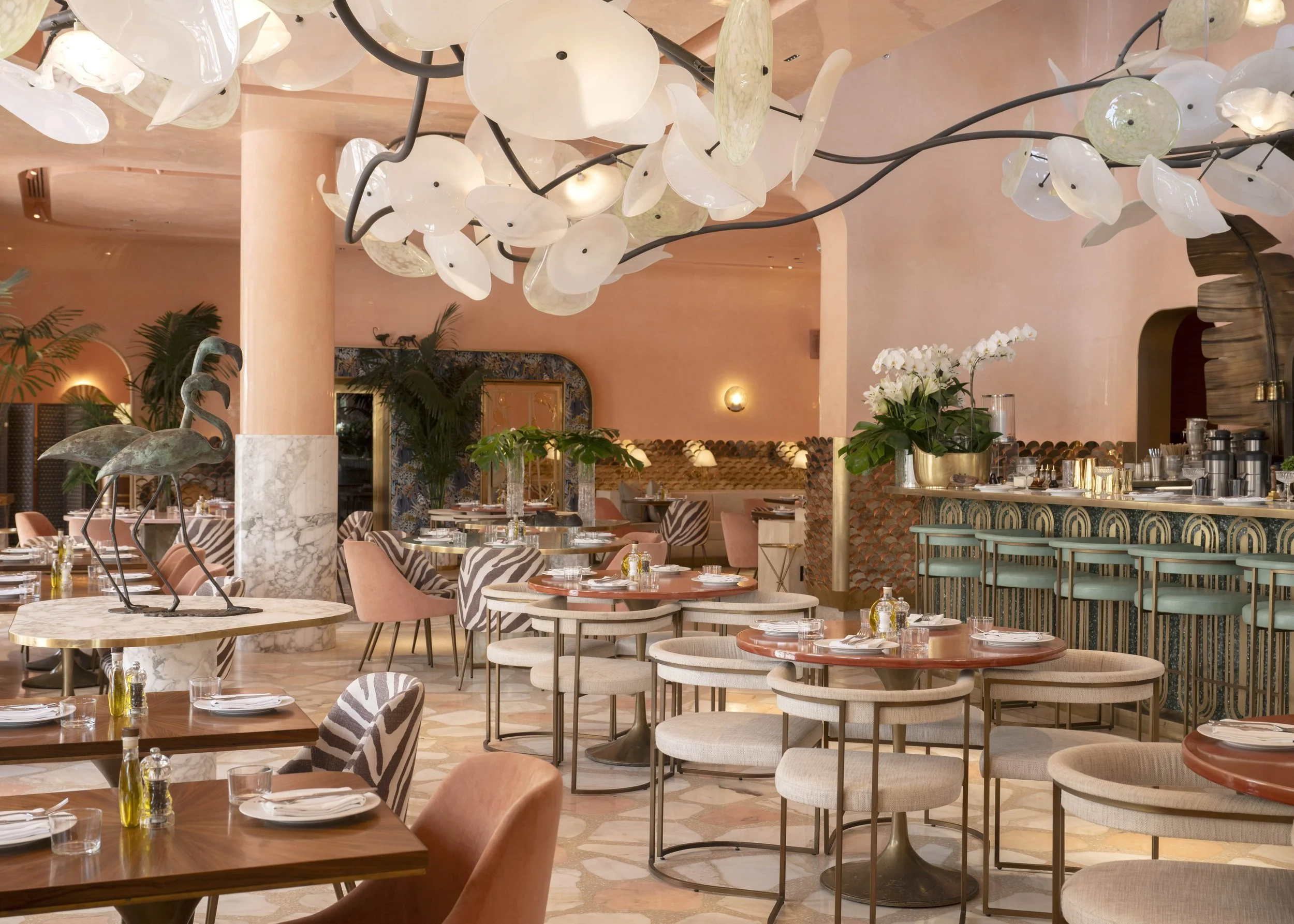 Elegant restaurant interior with pastel pink walls, marble accents, a large floral chandelier, and a bar with tiered seating, glassware, and decorative plants.