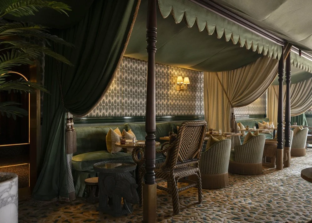 An elegant restaurant interior with dark green canopy drapes, patterned wallpaper, and a mix of upholstered chairs and a green velvet banquette seating. Table settings include napkins and glasses, and there is warm ambient lighting from wall sconces.