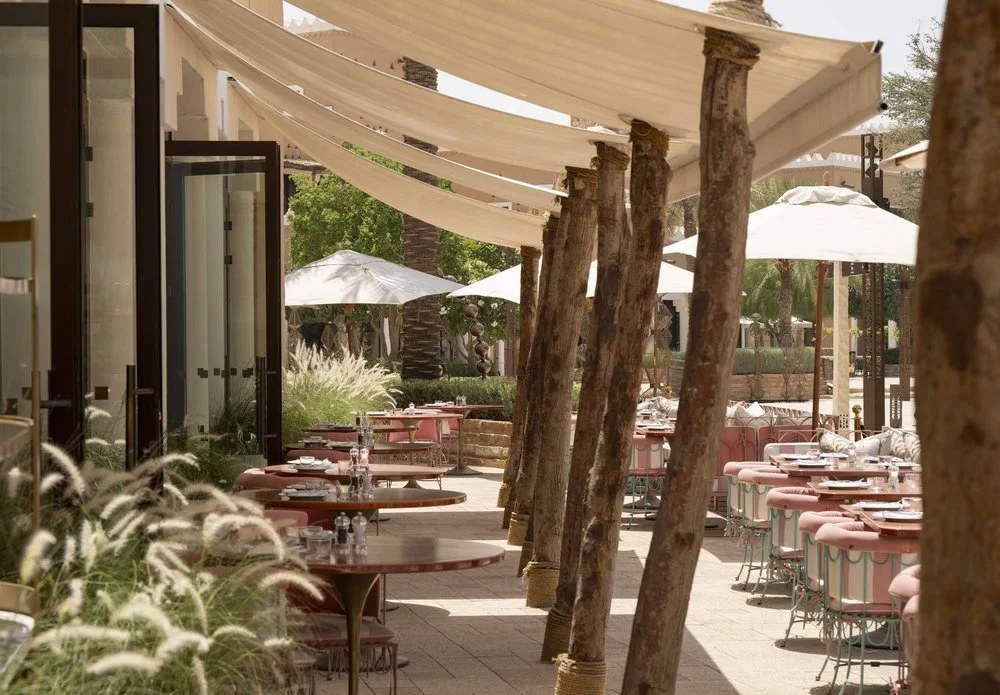 Outdoor restaurant patio with pink chairs, tables set with plates and glasses, white umbrellas, and a beige canopy overhead, surrounded by greenery and trees.