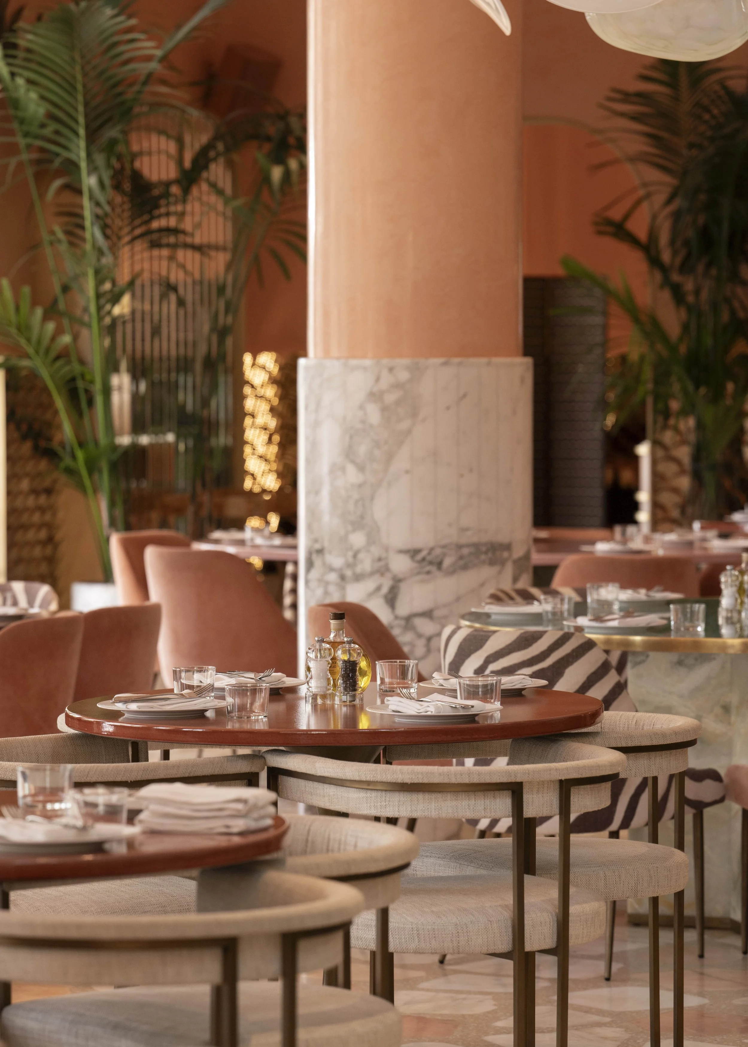 A stylish restaurant interior featuring multiple tables set with plates, glasses, and utensils. The decor includes large leafy plants, marble accents, and plush seating in neutral and earthy tones.
