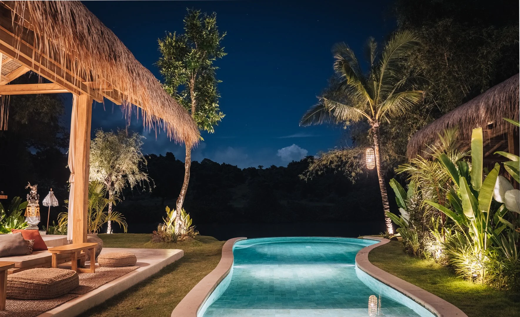 Nighttime scene of a tropical backyard with a swimming pool, illuminated by outdoor lights, surrounded by lush plants, palm trees, and thatched-roof structures.