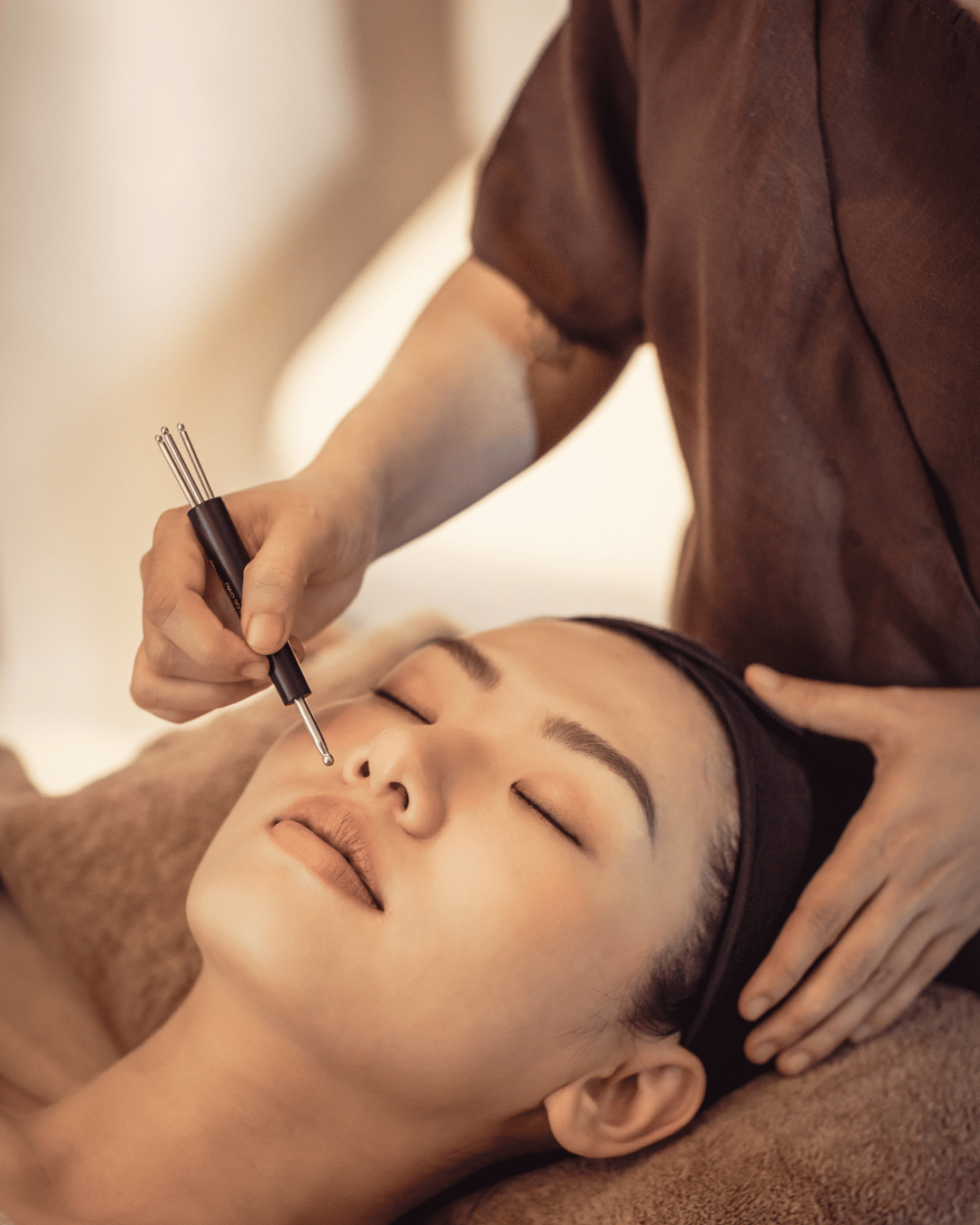 A woman receives a facial treatment from a professional using a small device, with her eyes closed and lying on a treatment bed. | Awakened Soul - Jessika Harrison 